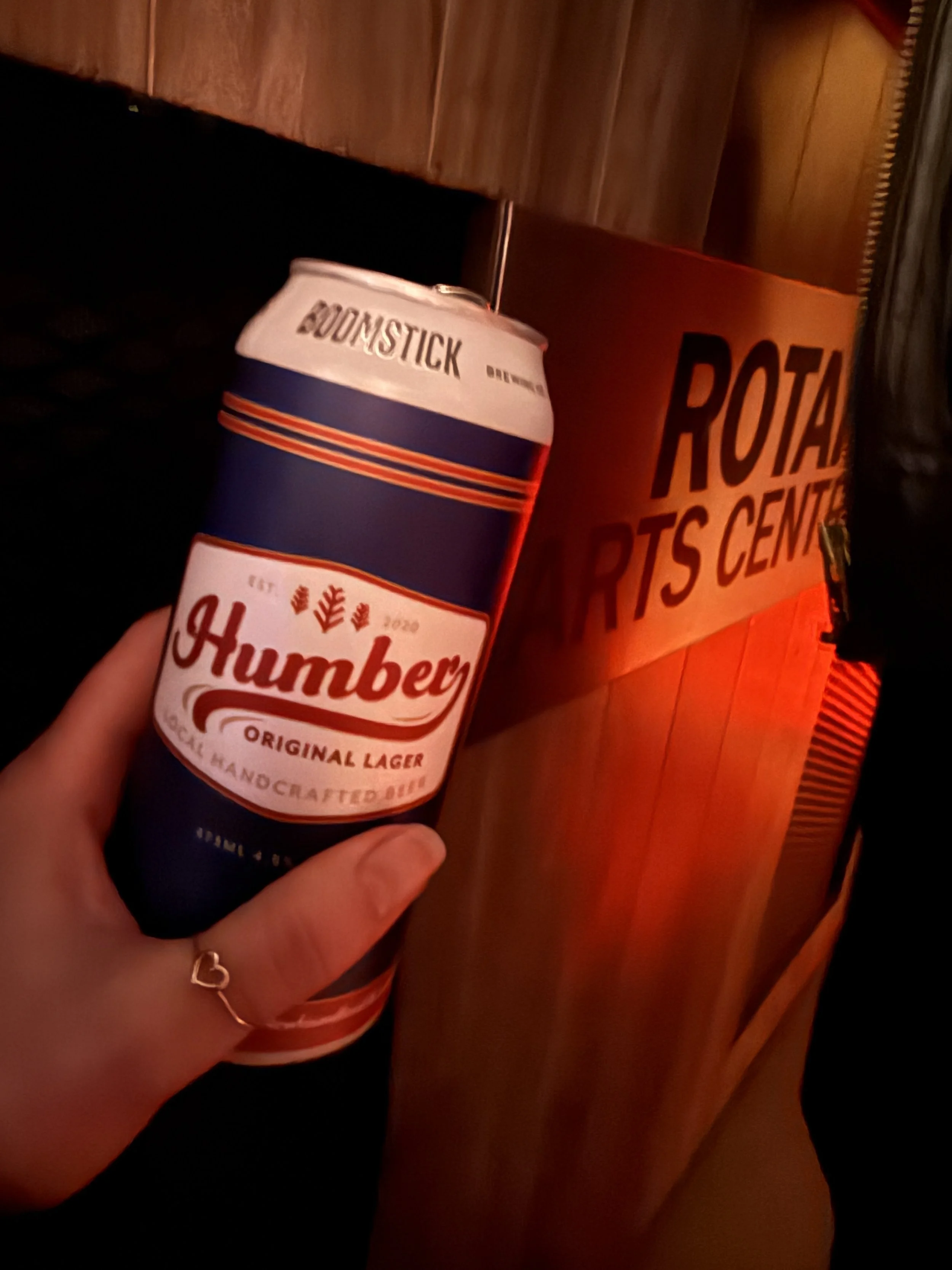 Person holding a can of Humburger beer in front of a wooden wall and a sign that reads "ROYA RTS CENTER".