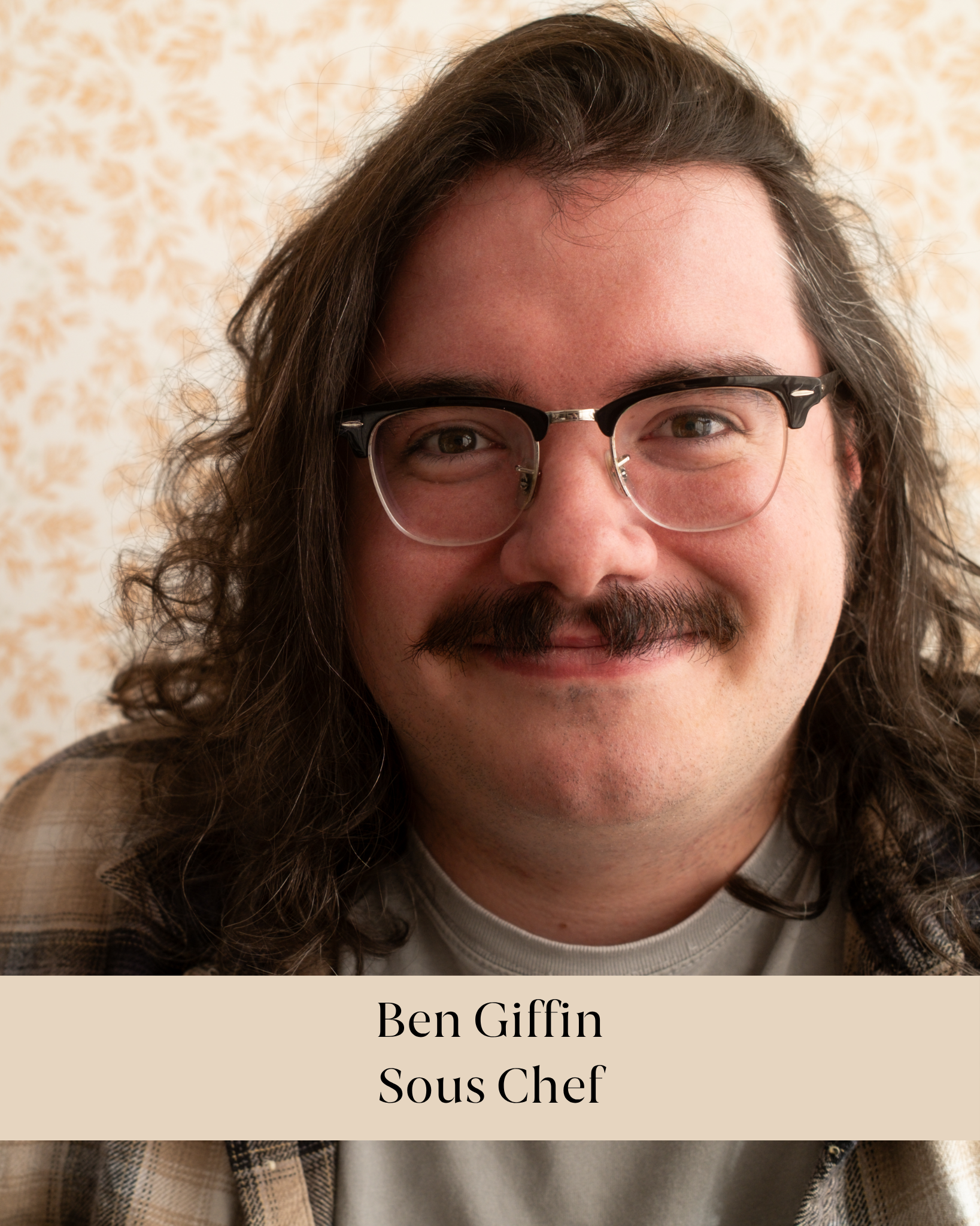 Close-up of a smiling man with long curly brown hair, wearing glasses and a beige plaid shirt, against a beige floral wallpaper background. Text overlay: "Ben Giffin Sous Chef".