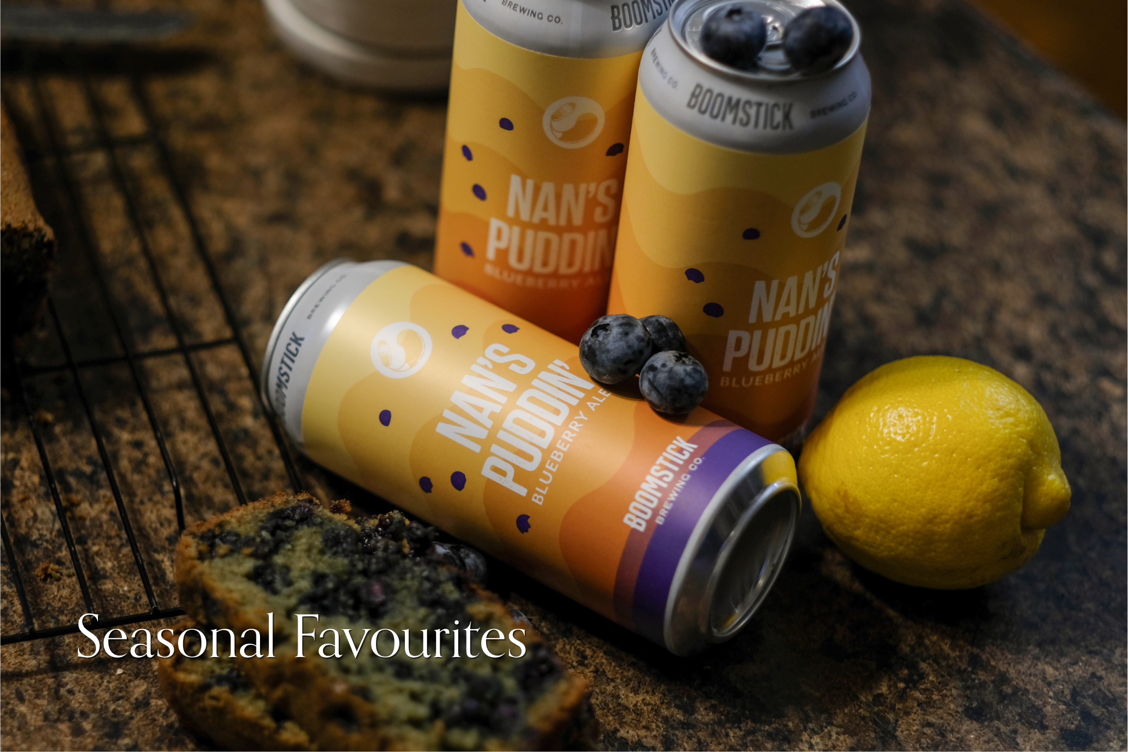 Three cans of Boomstick Brewing Co.'s Nan's Pudding blueberry ale on a dark countertop, with a lemon and blueberry, and a blueberry muffin nearby.