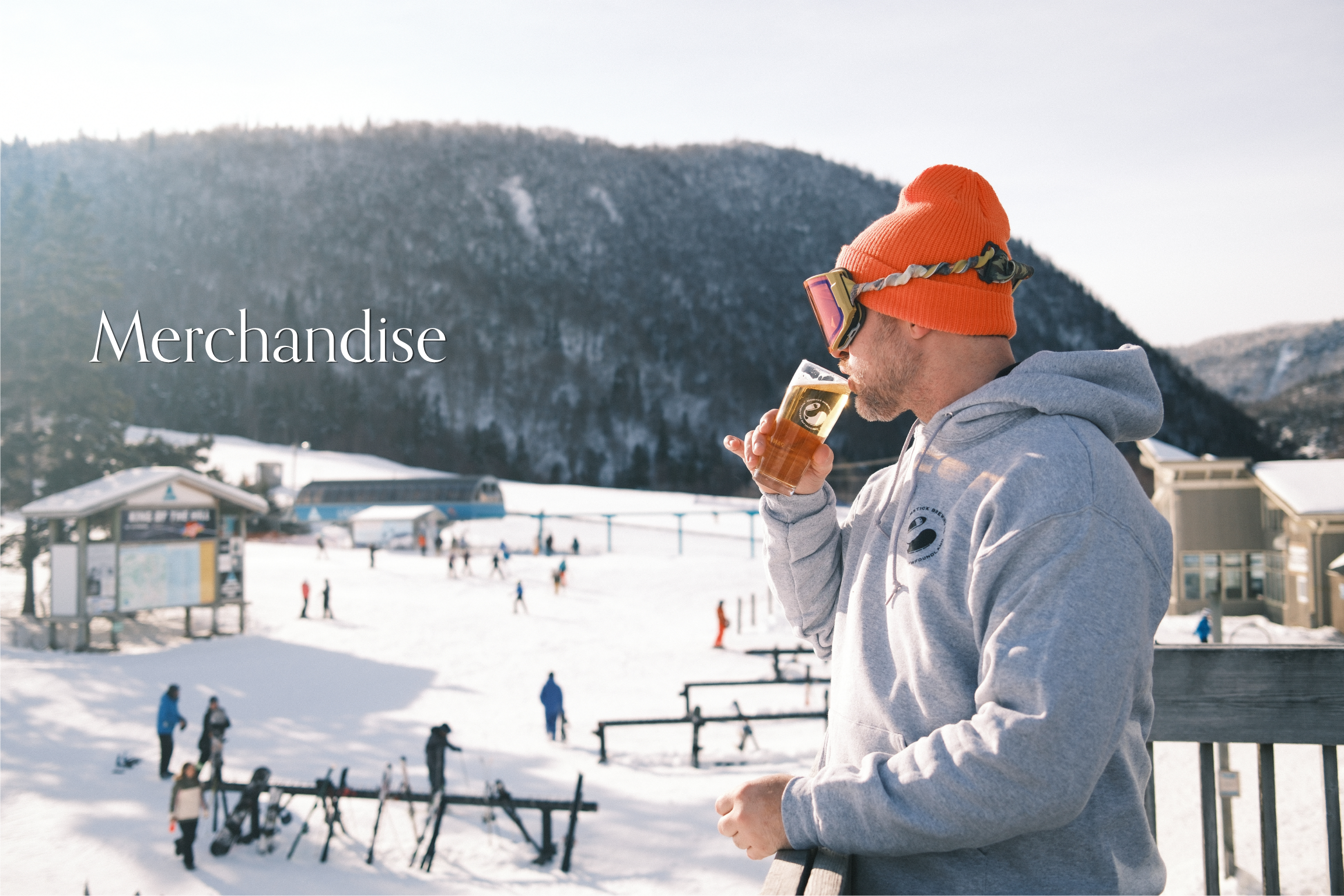 A man in a gray hoodie and orange beanie drinking beer on a snowy ski resort with skiers and snowy hills in the background.