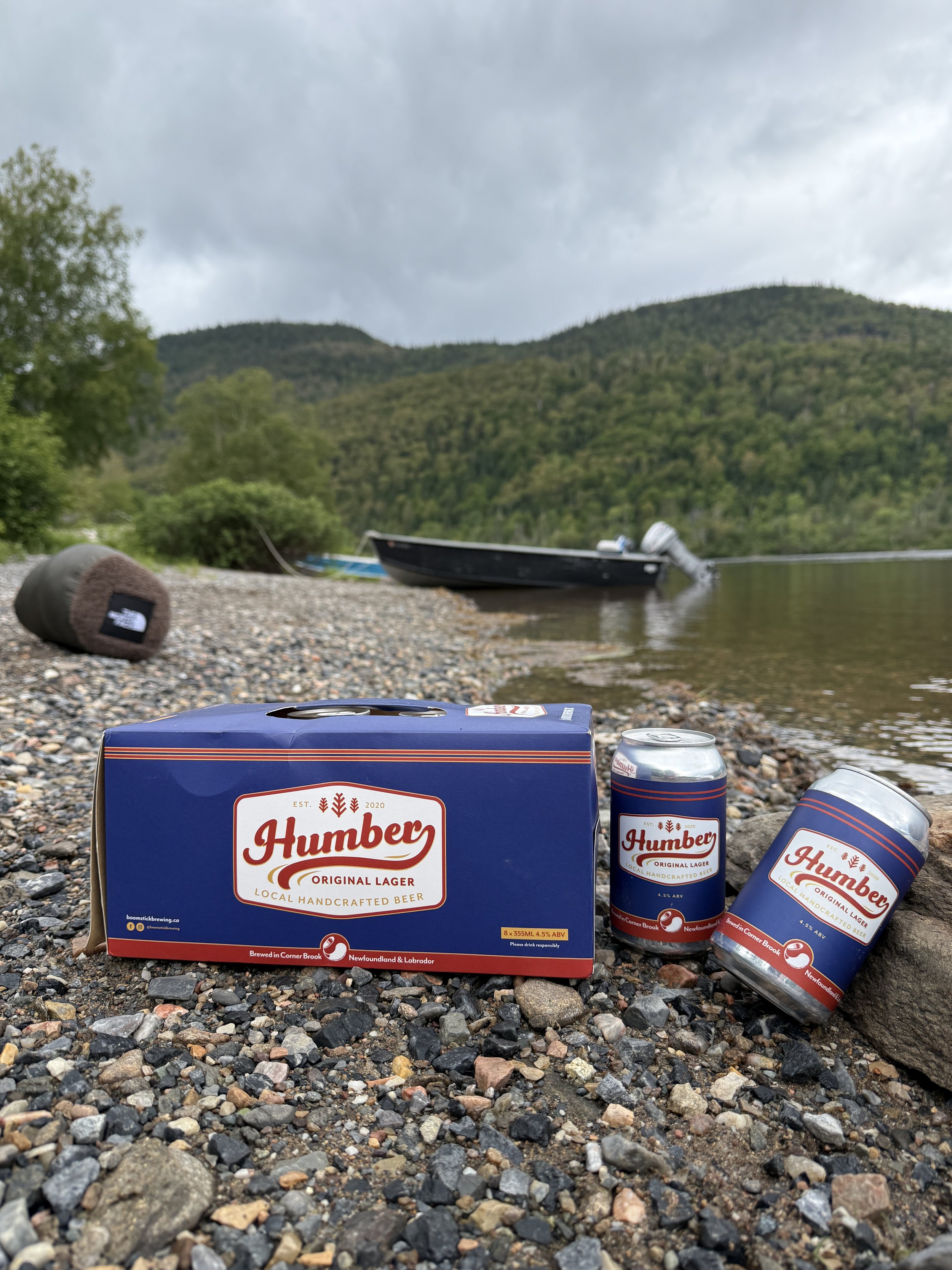 A box of Humbery Original Lager beer and three cans of the same beer on a pebbly beach near water with boats, trees, and mountains in the background.