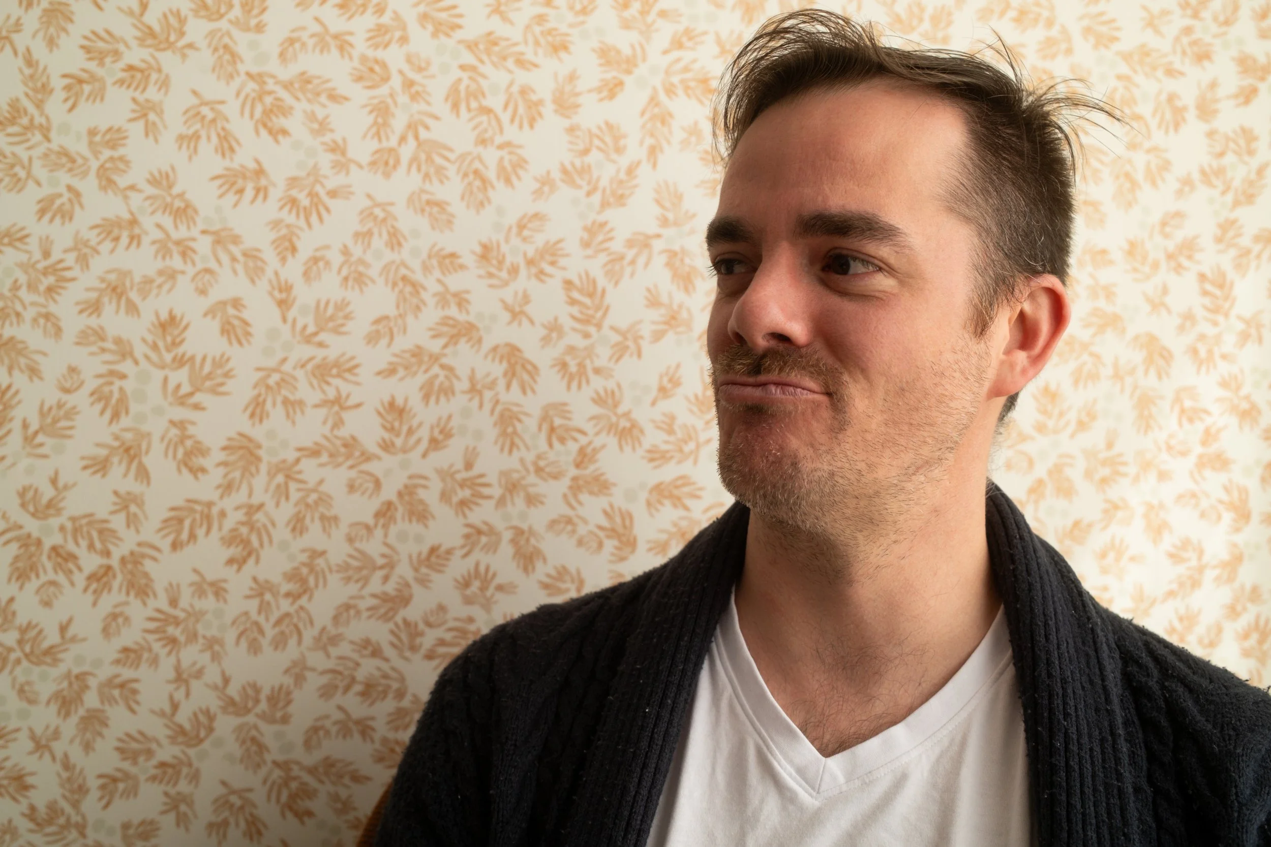 Close-up of a man with short brown hair and a beard, wearing a white shirt and black cardigan, standing in front of a beige patterned wallpaper.