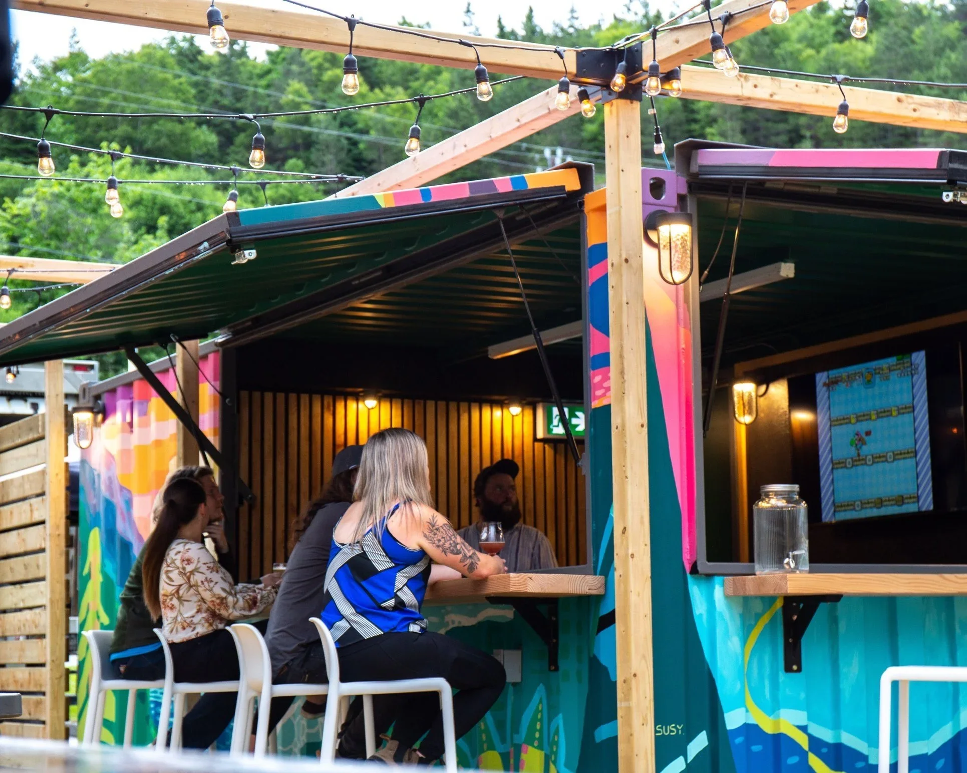 People sitting at a bar under a wooden structure decorated with string lights, with green trees in the background.