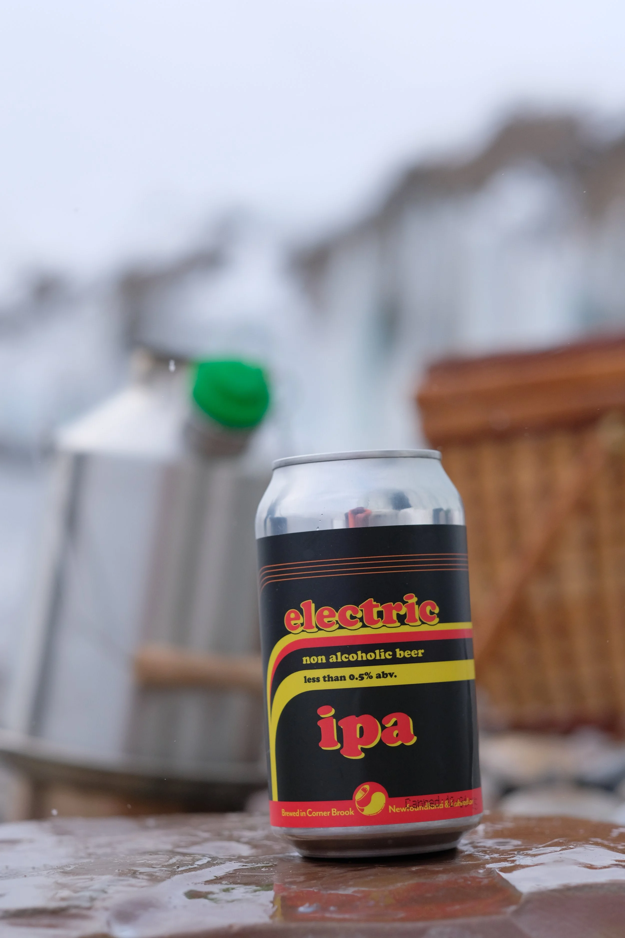 A can of non-alcoholic beer labeled 'electric' on a wet surface, with blurred objects in the background including a green cap and wooden structure.