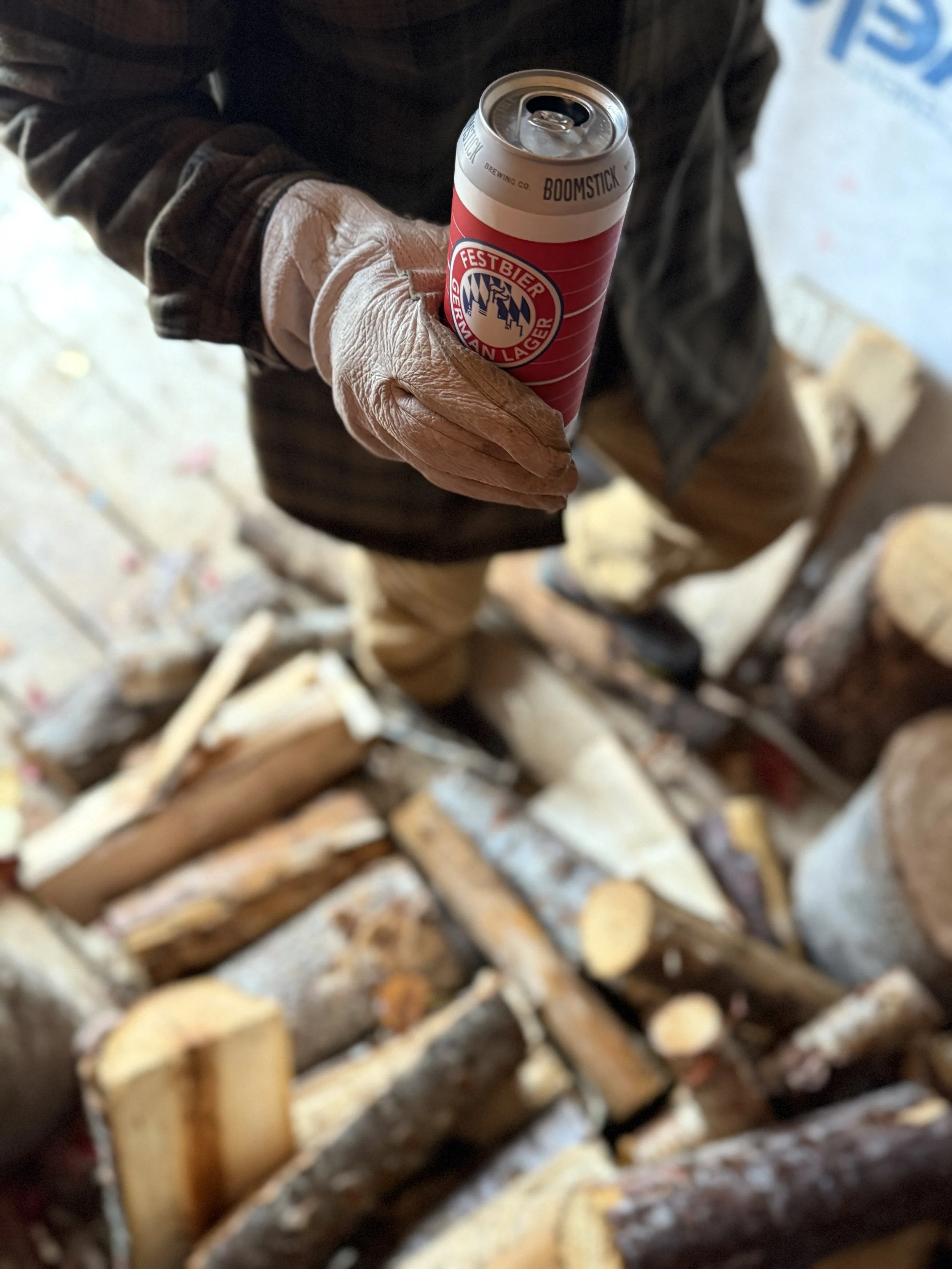 Person wearing a black jacket and brown leather glove holding a red and white canned beverage in front of a pile of chopped firewood.