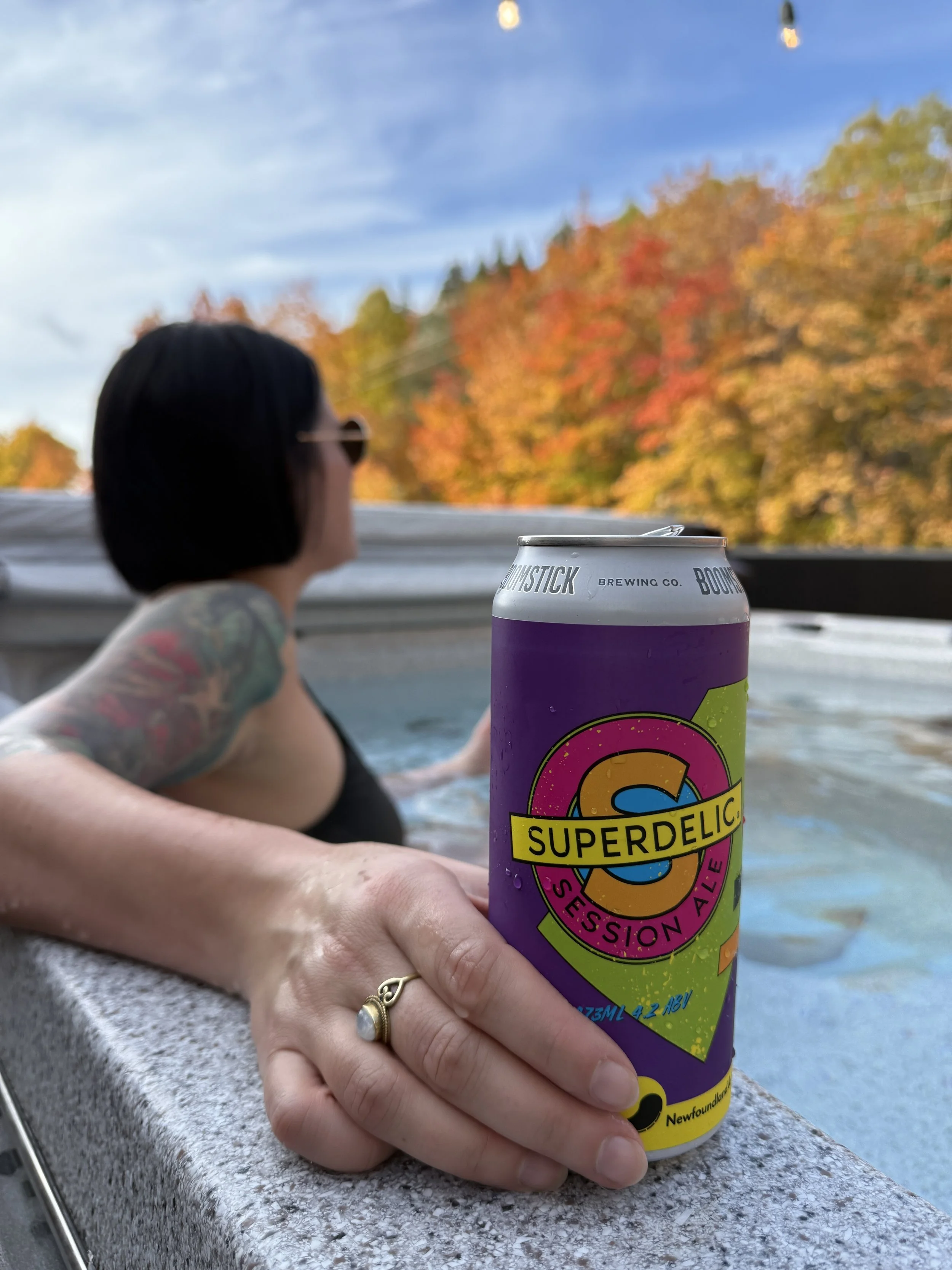 A person with tattooed arm and wearing a ring on finger is resting their hand on a concrete surface while holding a purple can of Super Delic session ale. In the background, a woman with dark hair and sunglasses is relaxing in a hot tub, with autumn-