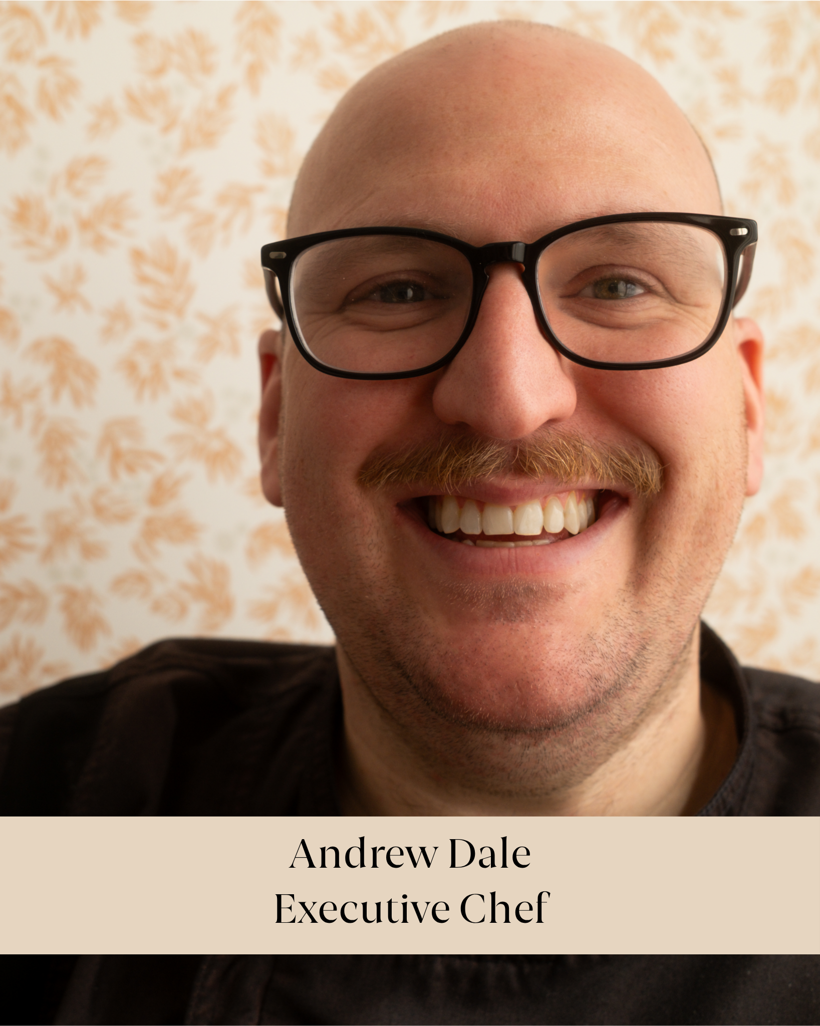 Close-up photo of a smiling man with glasses and a mustache, with a background of beige floral wallpaper. Part of a caption below identifies him as Andrew Dale, an executive chef.