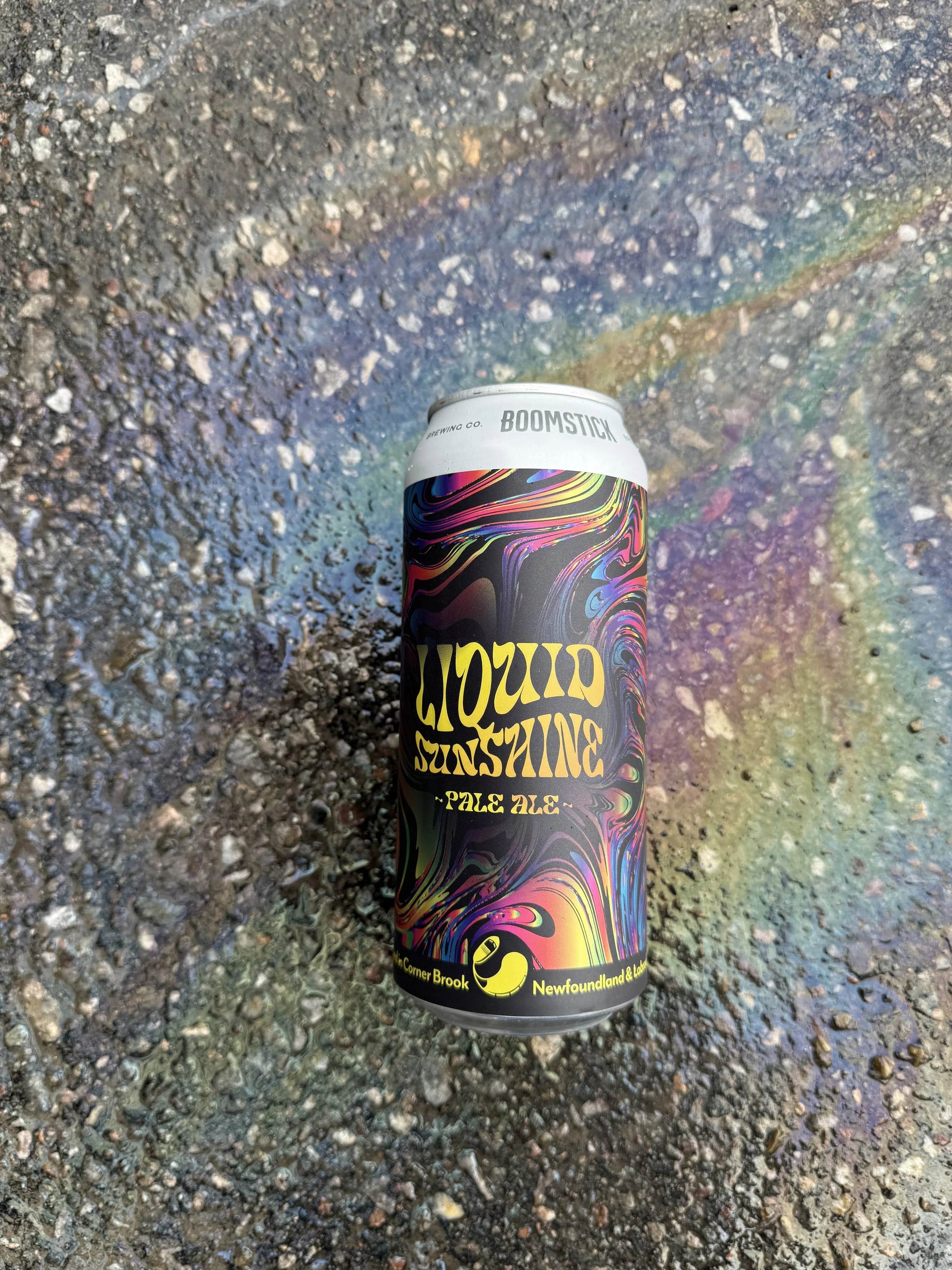 Colorful can of beer called 'BOOMSTICK' with psychedelic swirl design, sitting on rough asphalt ground, with rainbow-colored oil or residue around it.