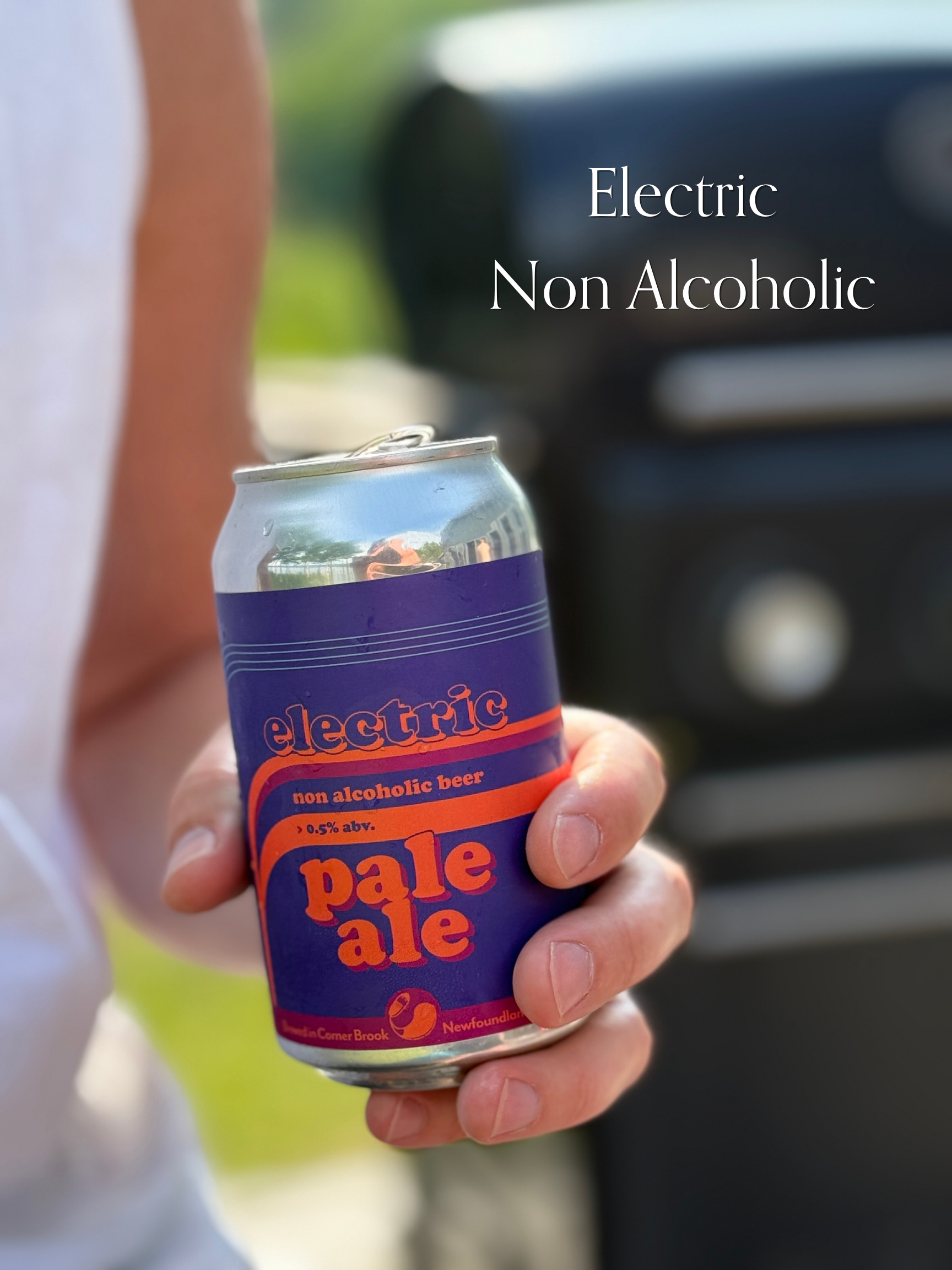 A person is holding a can of electric non-alcoholic beer called 'pale ale' in their hand. The can is purple and orange, and the background appears to be outdoors with blurred greenery and a car.