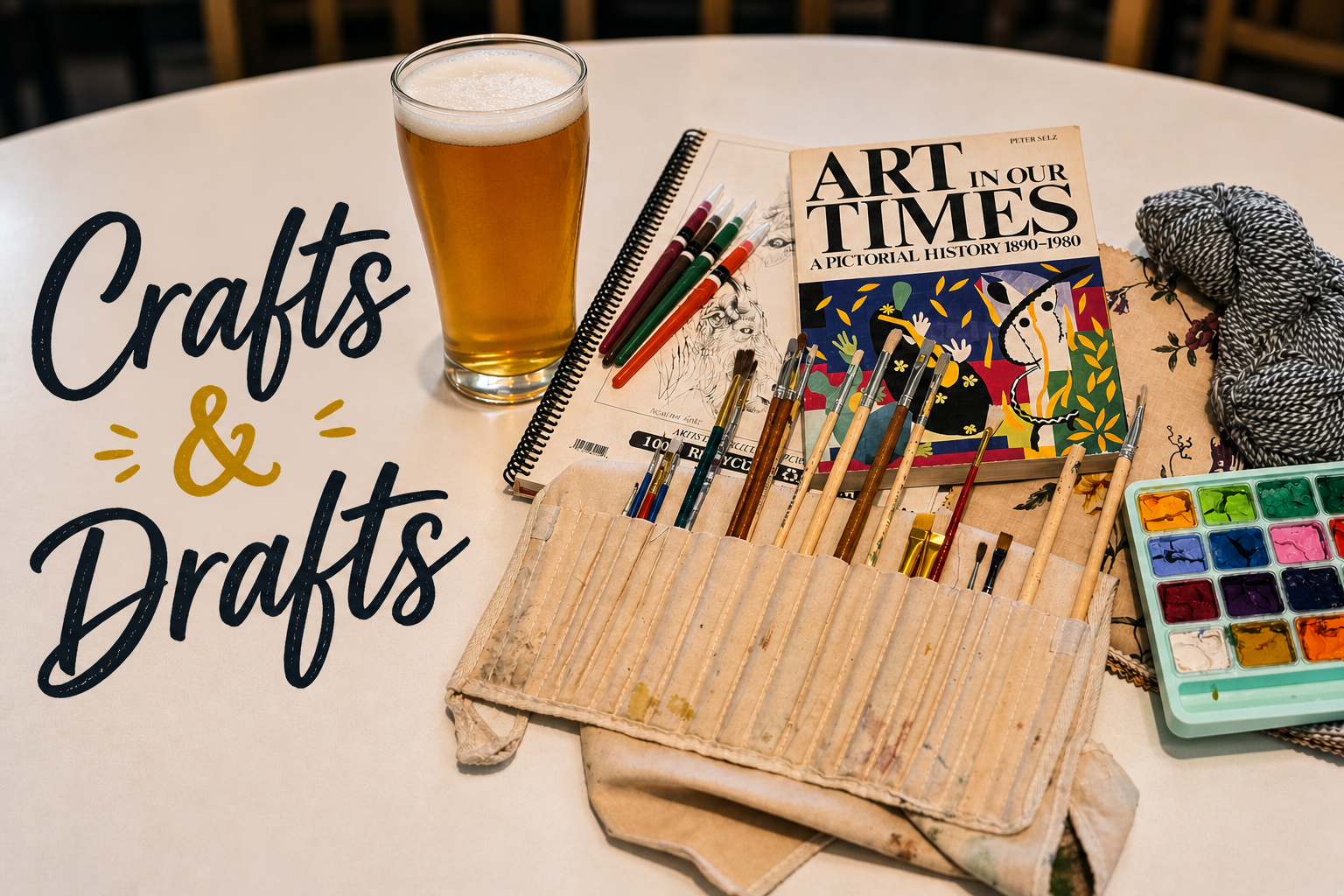 A table with art supplies, including watercolor paints, brushes, colored pencils, a notebook, a book titled 'Art in Our Times,' and a glass of beer, with the words 'Crafts & Drafts' written on the table.