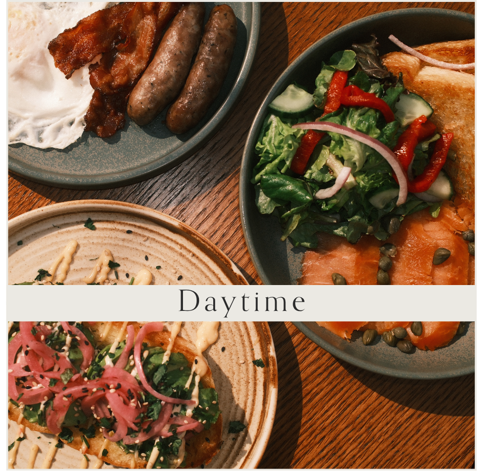 A top-down view of three plates of food on a wooden table. The plate in the top-left corner has two sausages, bacon, and a fried egg. The plate on the right contains a mixed green salad with cucumbers, tomatoes, red onion, and smoked salmon, garnishe