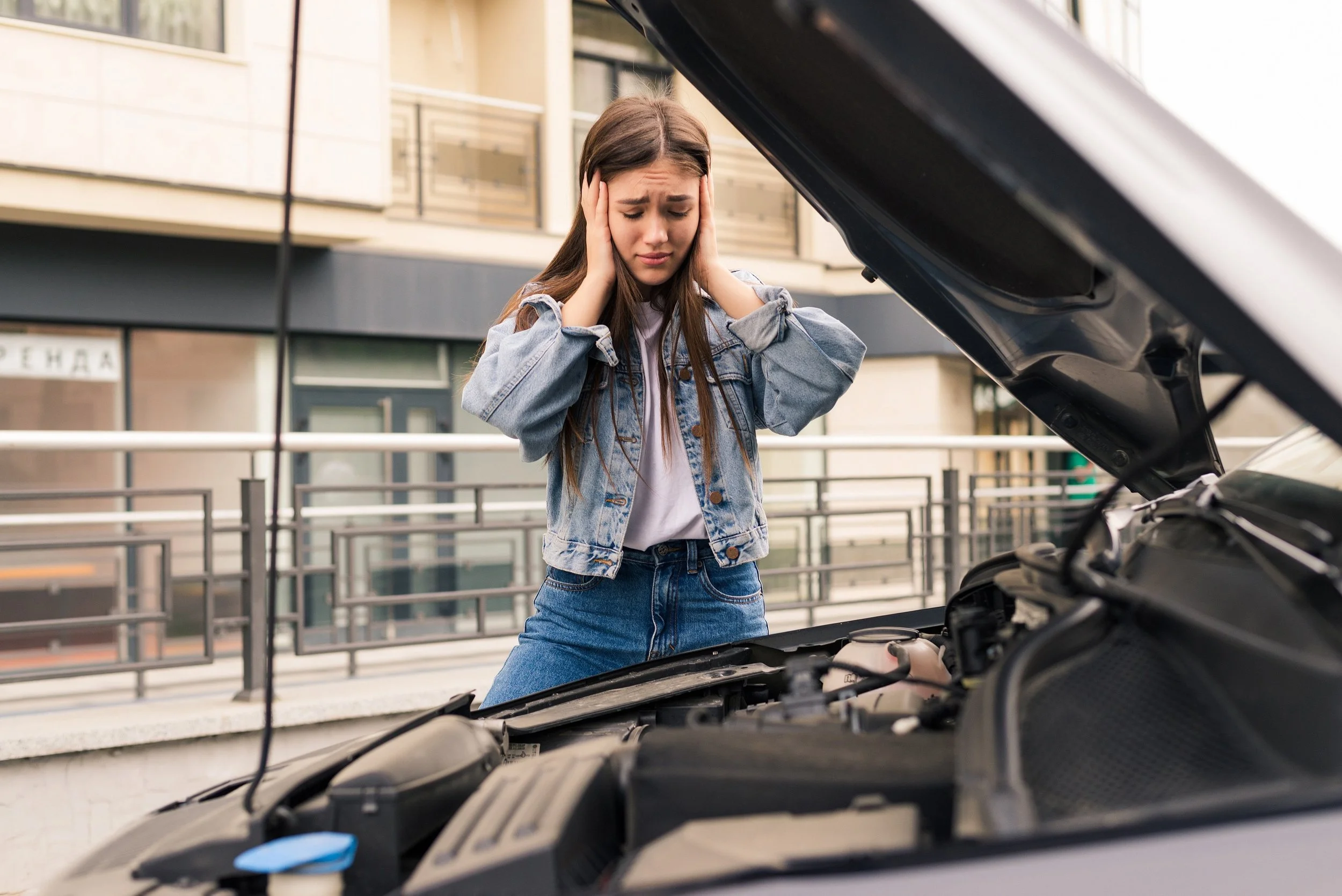 A young woman with long brown hair wearing a denim jacket and jeans looking worried while holding her head with both hands, standing in front of an open car hood, on a city street with buildings and railings in the background.