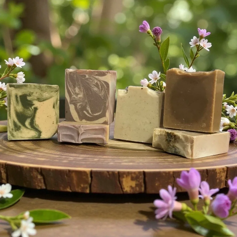 A variety of handmade soap bars with marbled and solid patterns, arranged on a wooden round platter surrounded by small purple and white flowers and greenery.