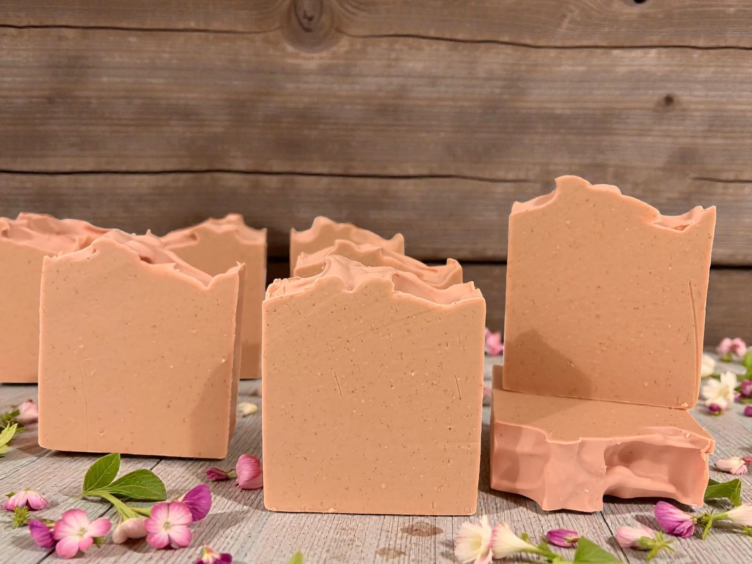 A group of pink soap bars with a wavy top edge arranged on a wooden surface, surrounded by pink and white flower petals.