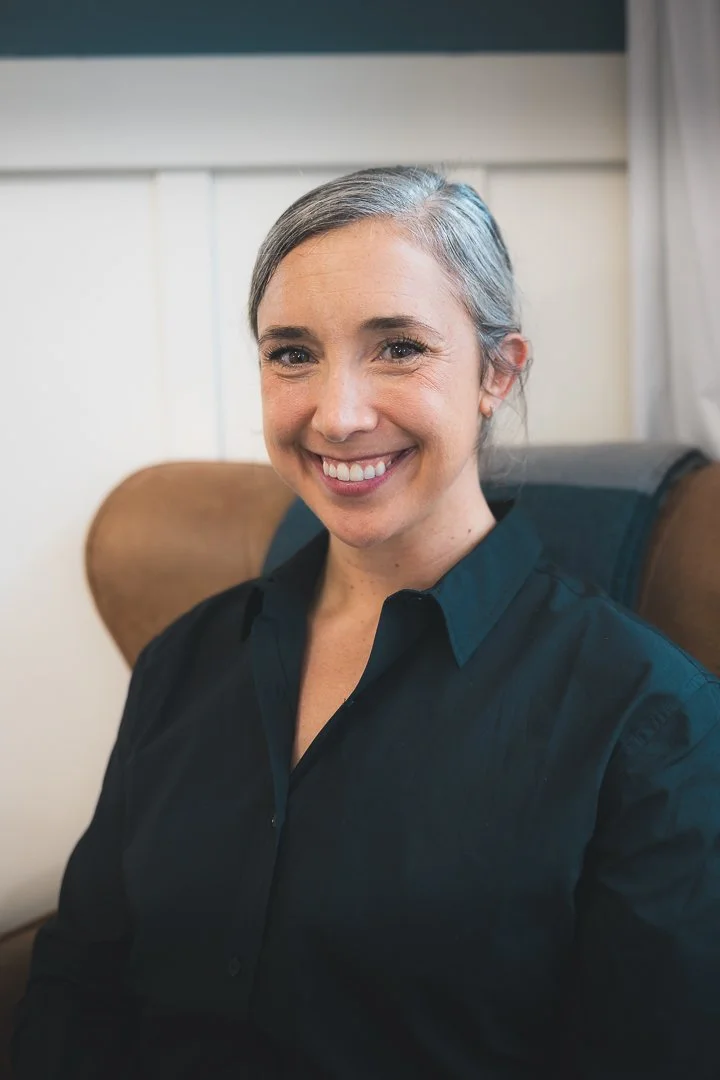 A smiling woman with gray hair tied back, wearing a dark button-up shirt, sitting in a brown chair in a room with white paneling and curtains.