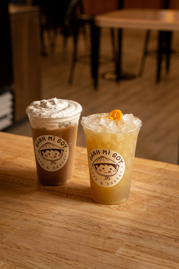 Two iced beverages in to-go cups with the Banh Mi Boy logo, one topped with whipped cream and the other with crushed ice and an orange garnish, on a wooden table in a cozy café setting.