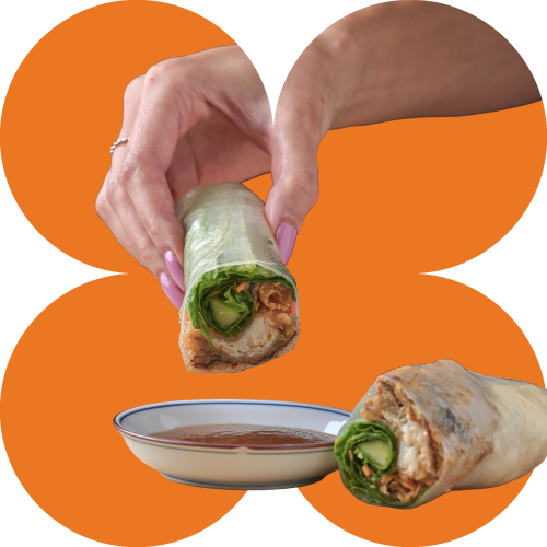 A hand holding a Banh Mi Boy spring roll dipped in sauce, with another spring roll on a plate in front, against an orange background.
