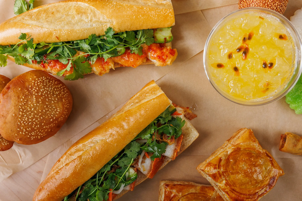Two Banh Mi Boy sandwiches with lettuce and chicken, a sesame seed bun, a bowl of mango passion fruit pudding, and pieces of pastry on a brown paper background.