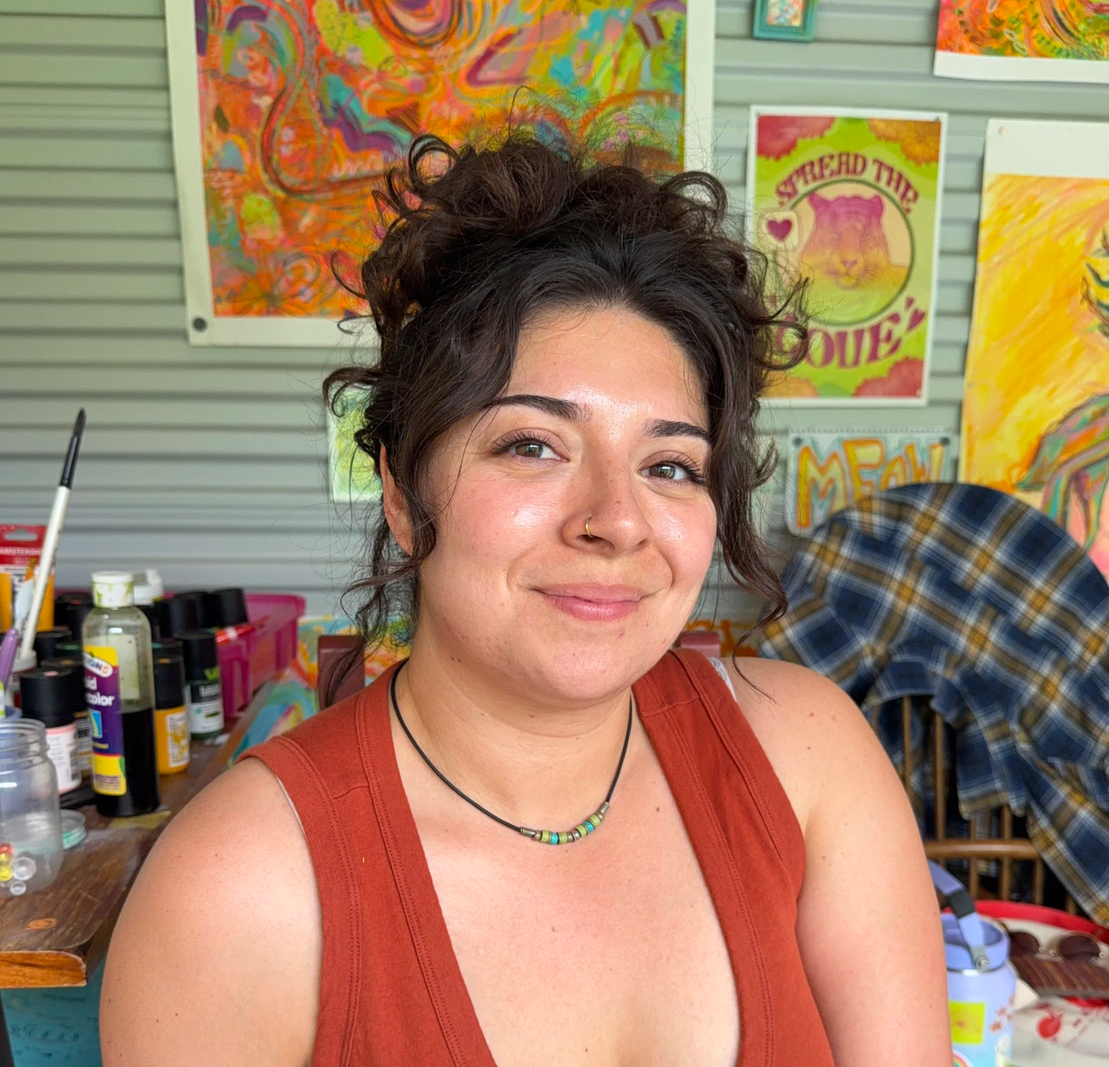 A young woman with curly dark hair, wearing a red tank top and a colorful bead necklace, smiling at the camera with colorful artwork and supplies in the background.