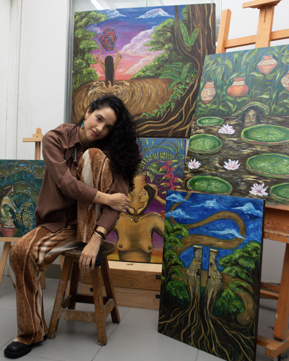 Portrait of Valeria R.O. seated in her studio, surrounded by paintings inspired by Amazonian landscapes, rivers, roots, and human figures.