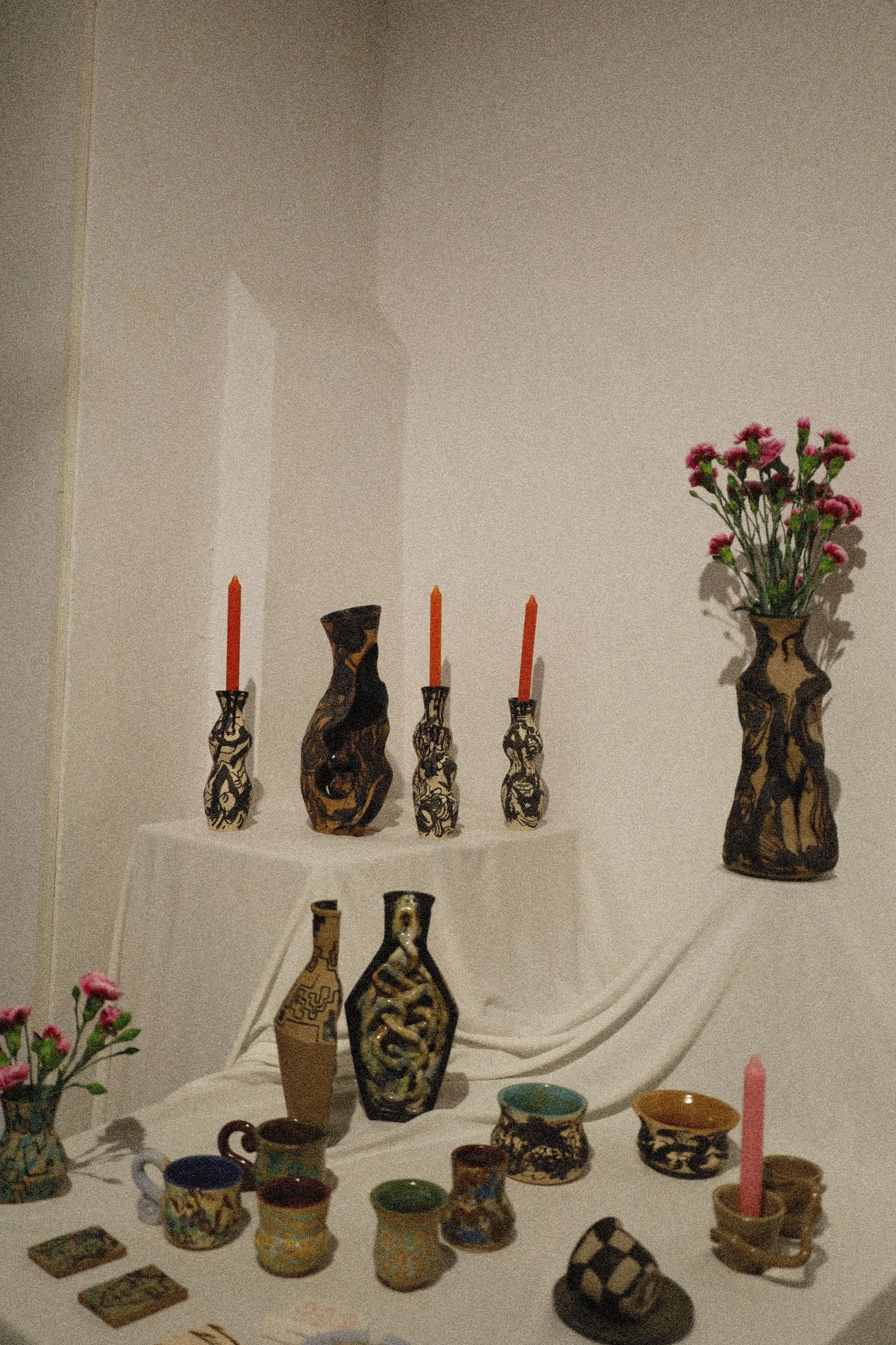 Vasos decorativos de cerámica con diseños en tonos marrón y negro, algunos con candeleros y velas naranjas, flores rosas en un jarrón grande, tazas y platos coloridos en una mesa.