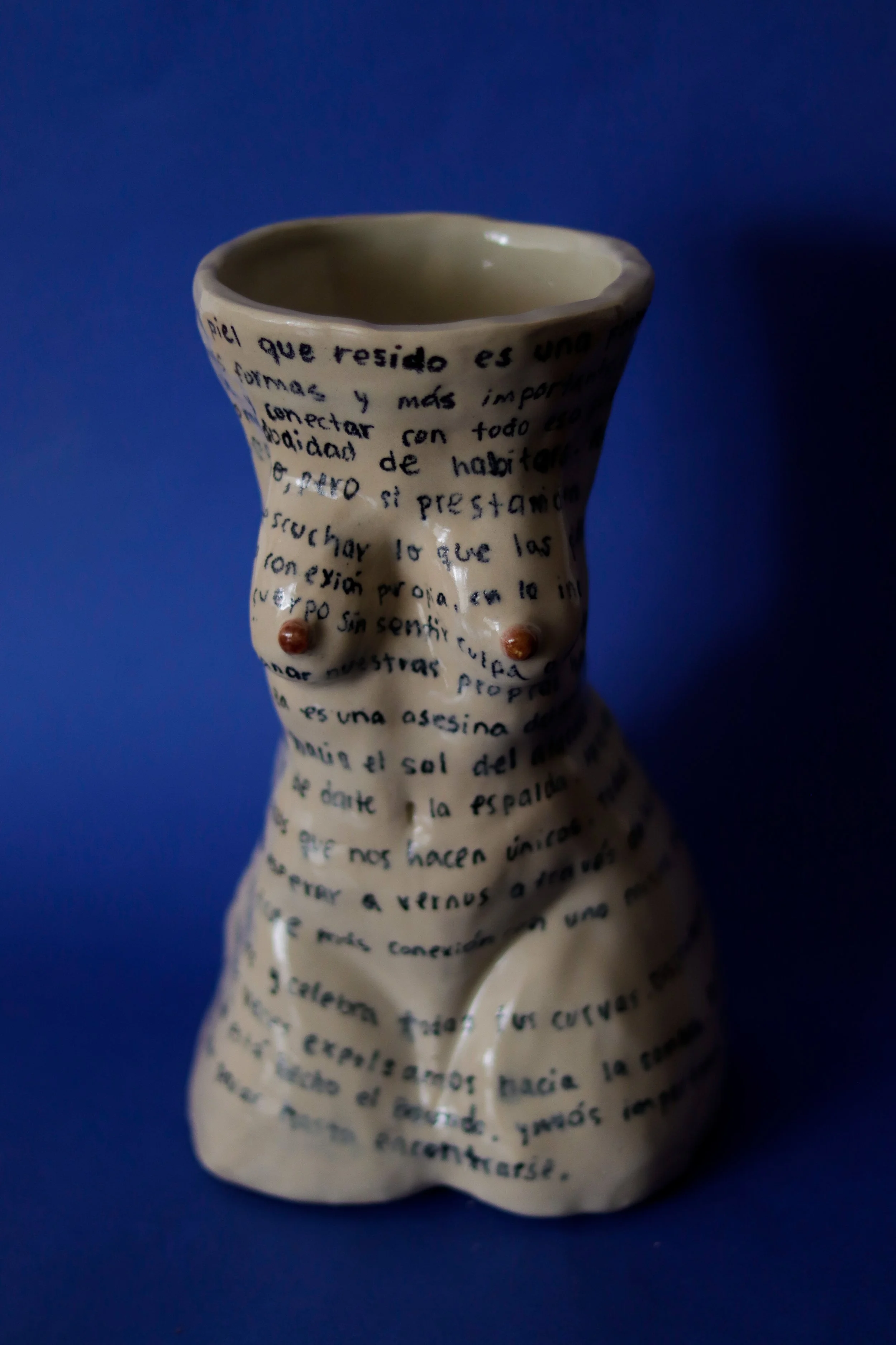 Sculptural ceramic vase shaped as a female figure, featuring handwritten inscriptions across its surface and photographed against a blue background.