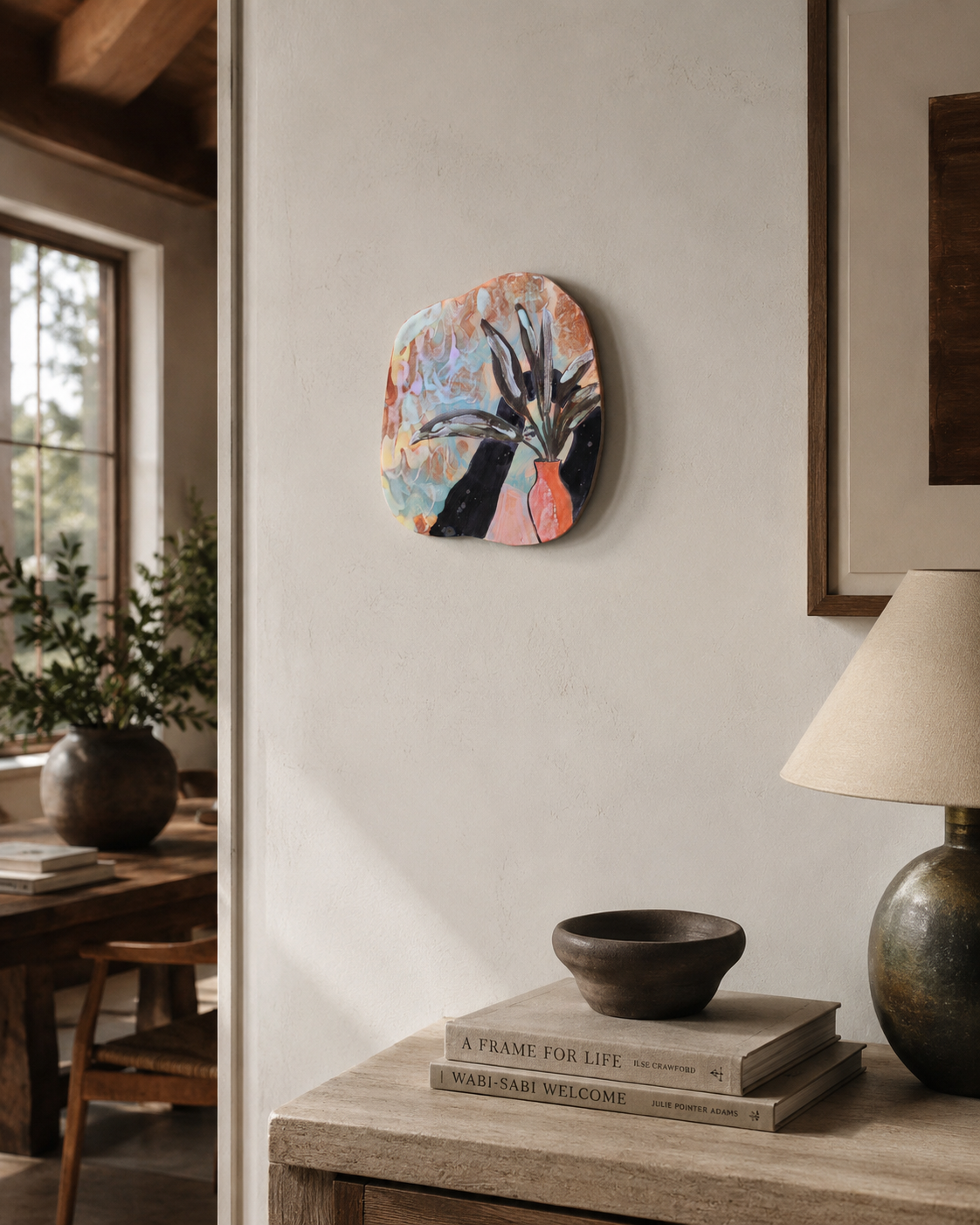 Hand-built ceramic wall piece displayed in an interior, featuring a botanical still life with dark leaves, a pink and orange vase, and layered pastel-colored glazes.