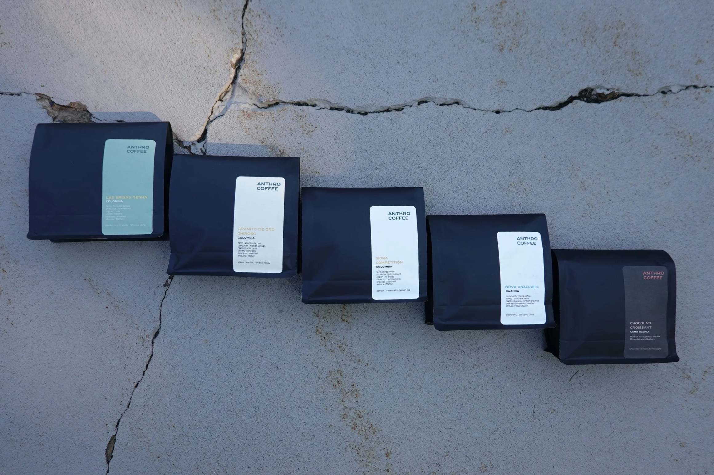 Four bags of coffee placed on a cracked concrete surface. The bags are black with labels indicating different coffee types from Anthro Coffee.