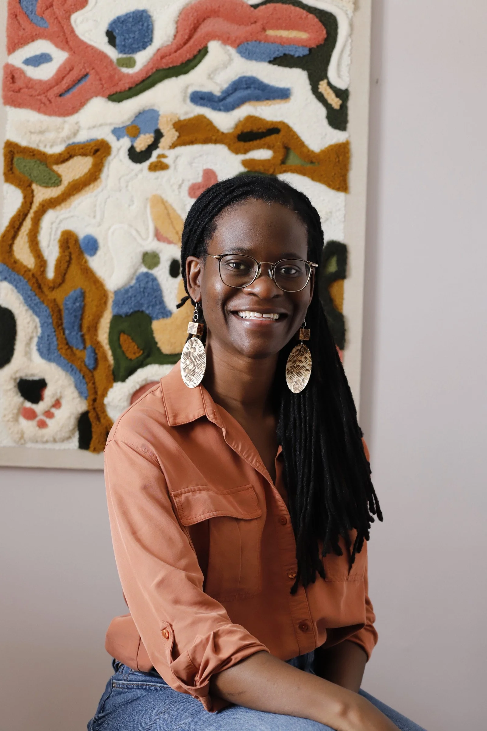 A woman with glasses and long black dreadlocks, smiling, sitting in front of a colorful abstract wall hanging.