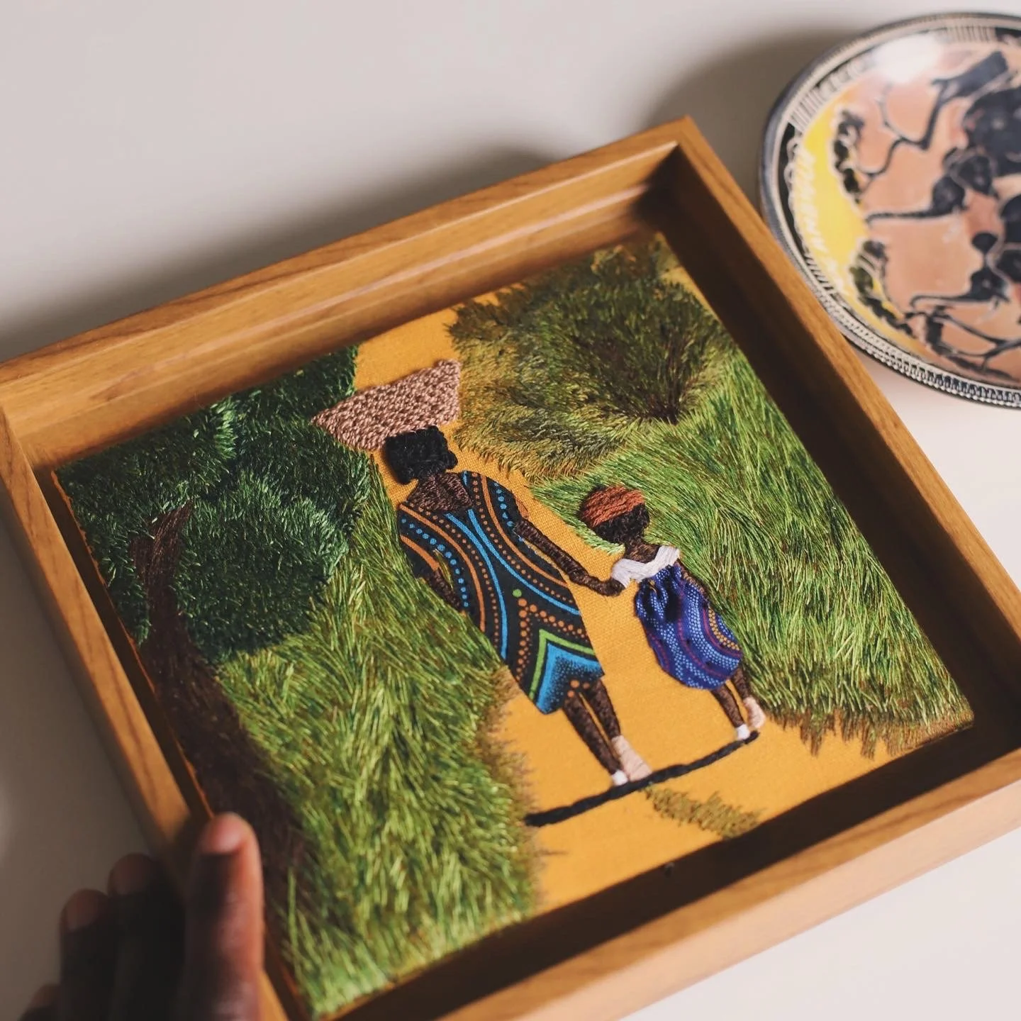 Embroidery artwork of two African children walking on a dirt path surrounded by greenery, with a wooden frame.