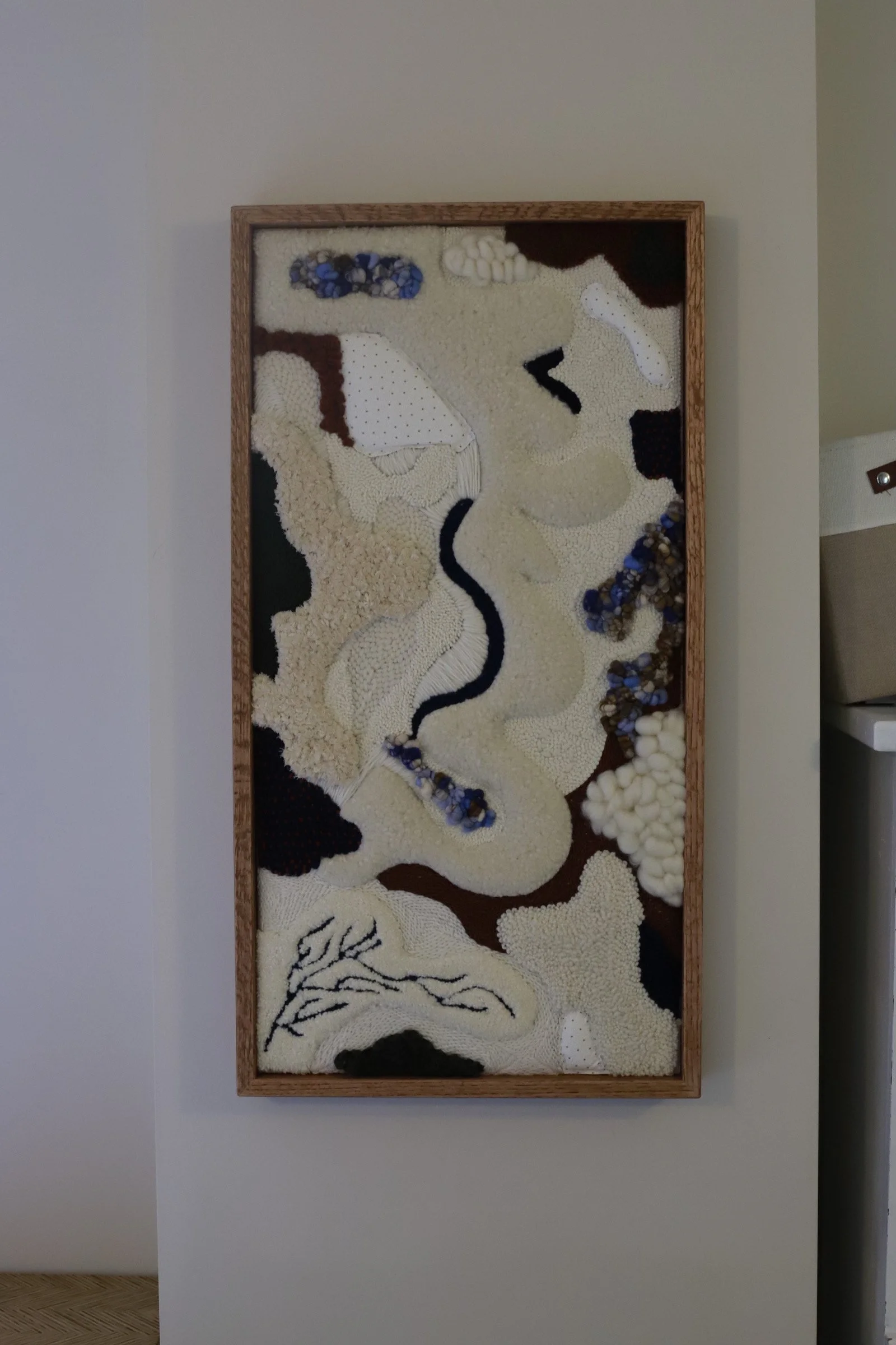 Mixed-media abstract art piece framed on a white wall, featuring textured materials in beige, brown, black, white, and blue, with organic and flowing shapes.