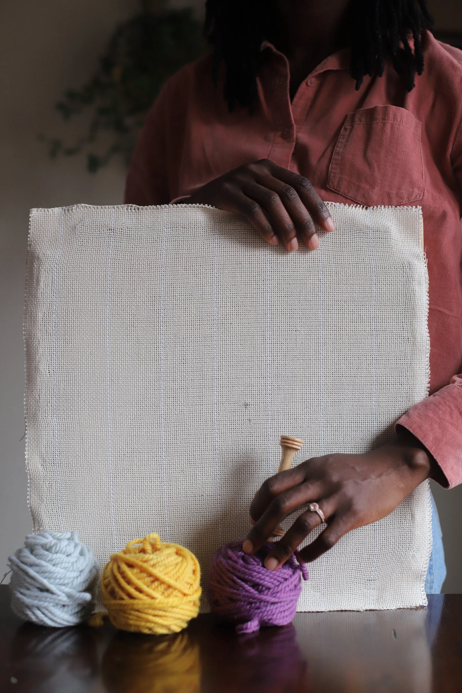 Person with dark skin, wearing a pink shirt, holding a blank beige canvas with yarn balls in white, yellow, and purple on a dark wooden table.
