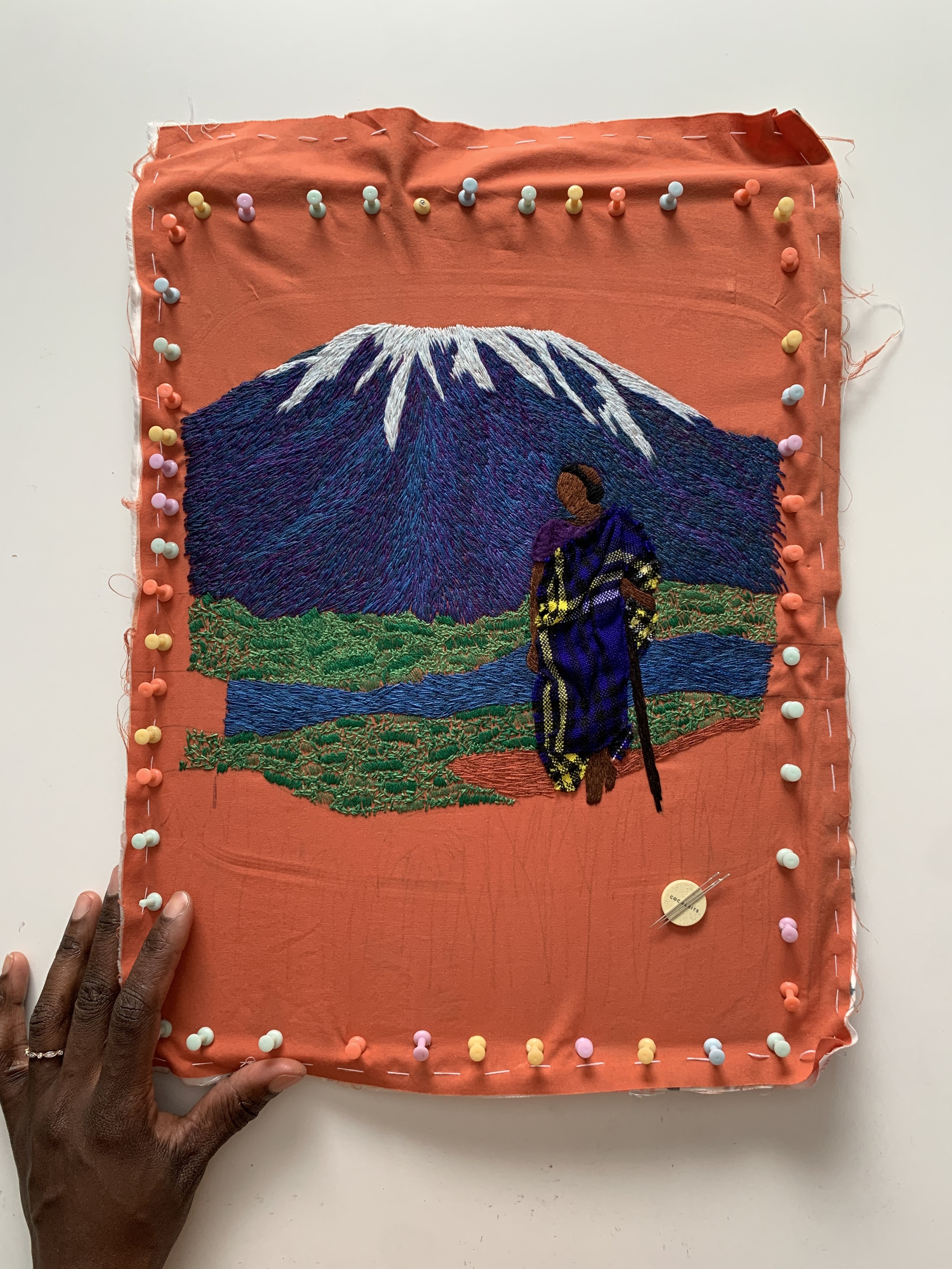 Embroidery artwork depicting a person in traditional clothing standing in front of a mountain with snow on its peak, green fields, and a river, on a pink fabric background with colorful pins around the border.