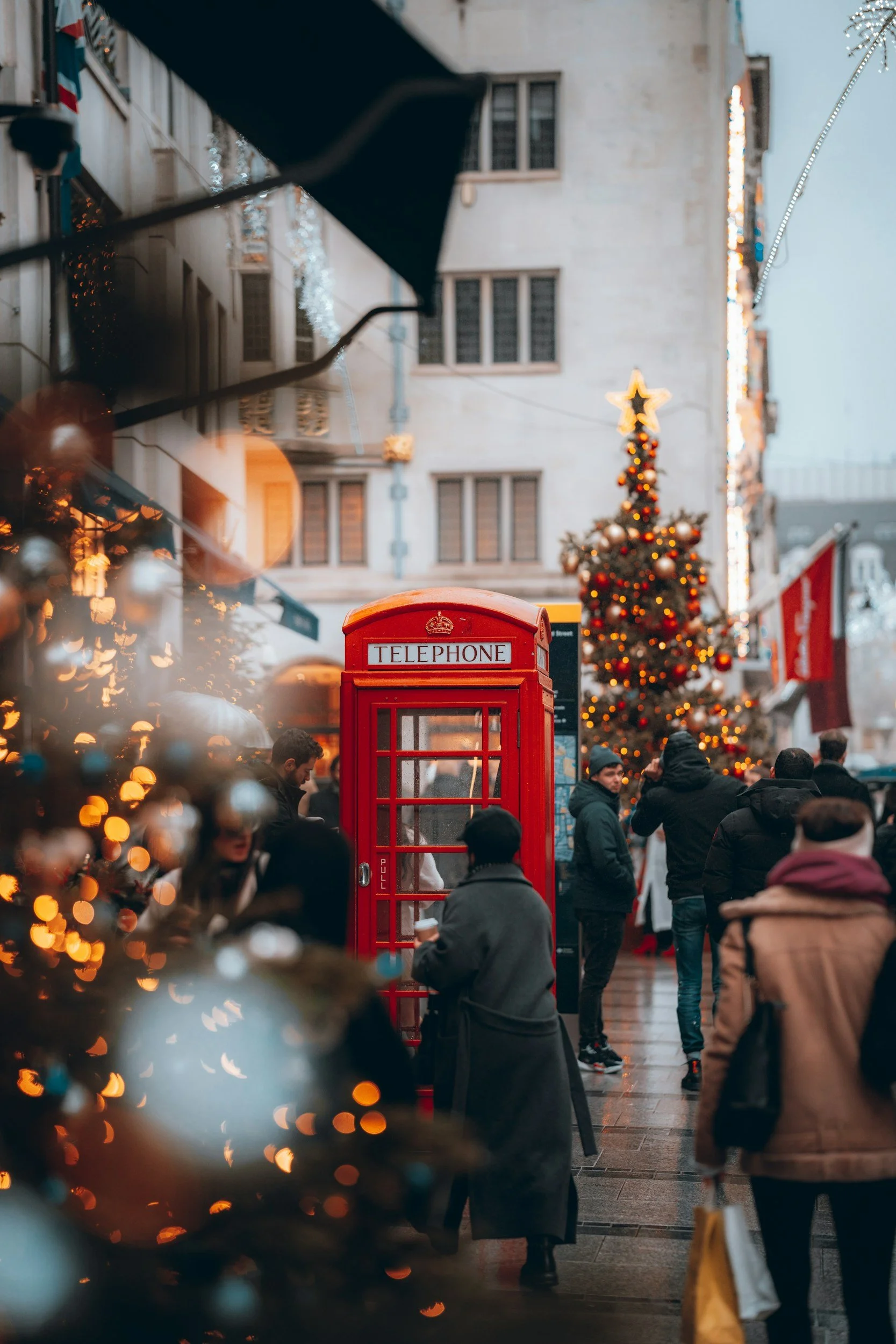A bustling city street decorated for Christmas with a large Christmas tree adorned with ornaments and lights, a red telephone booth, and people walking and gathering near. There are Christmas lights hanging across the street and buildings in the background.