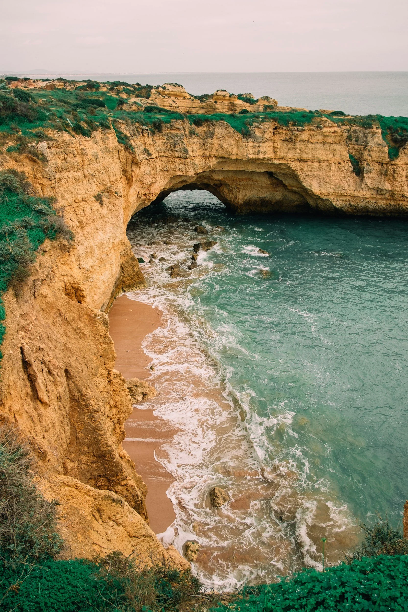 Cliff side view of a natural rock arch over a sandy beach with ocean waves, greenery on top, and cloudy sky in the background.