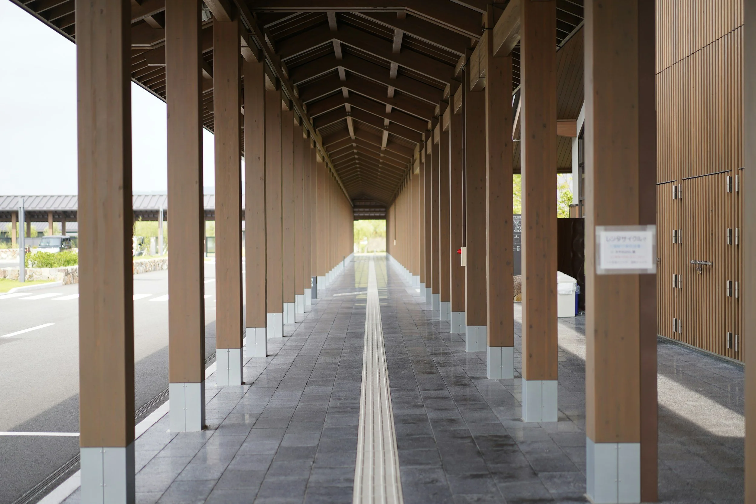Wooden walkway or passageway with a gabled roof, supported by vertical wooden beams, leading to an outdoor area with parking and greenery in the background.