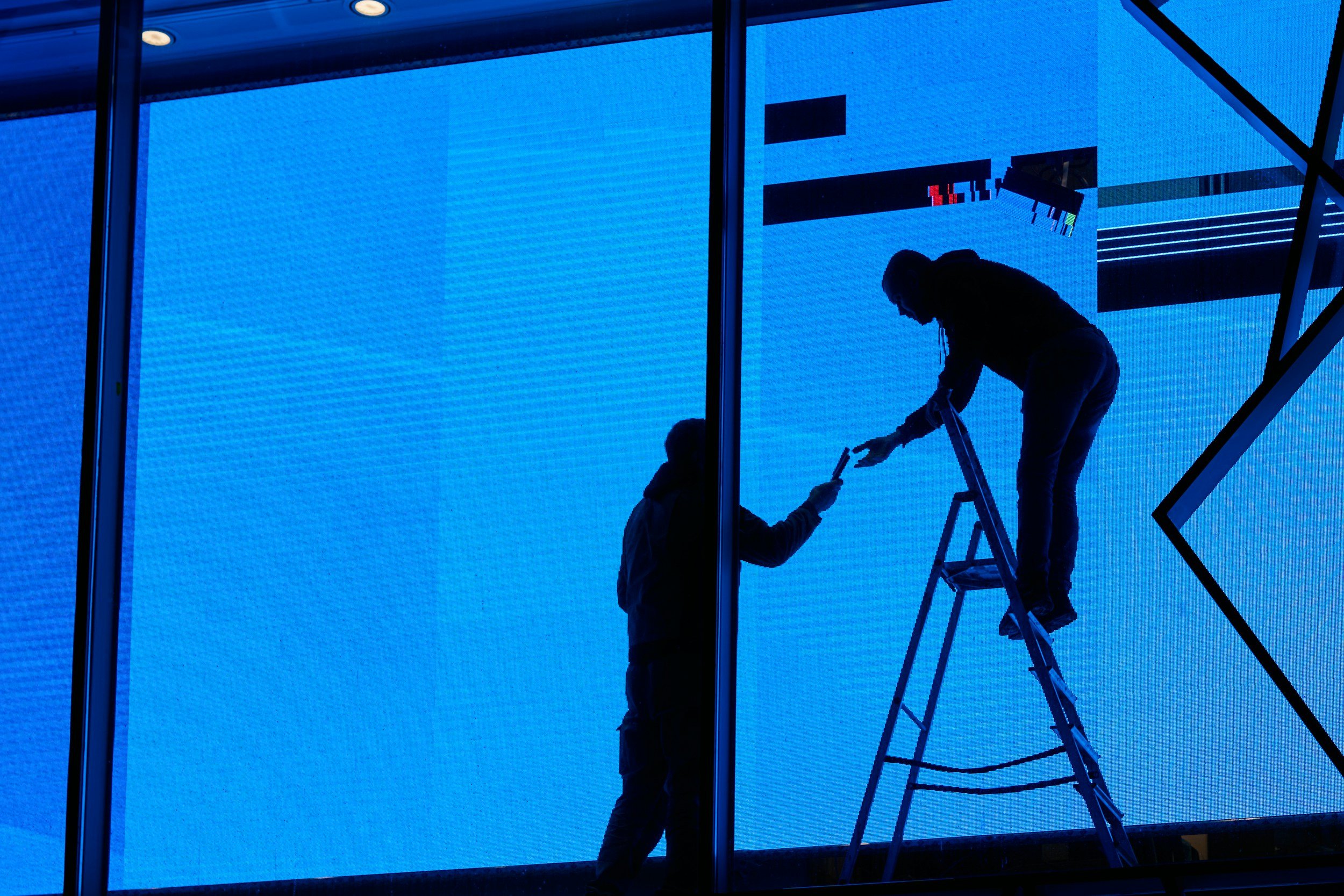 Silhouettes of two workers on a ladder working on a large digital screen with blue background.