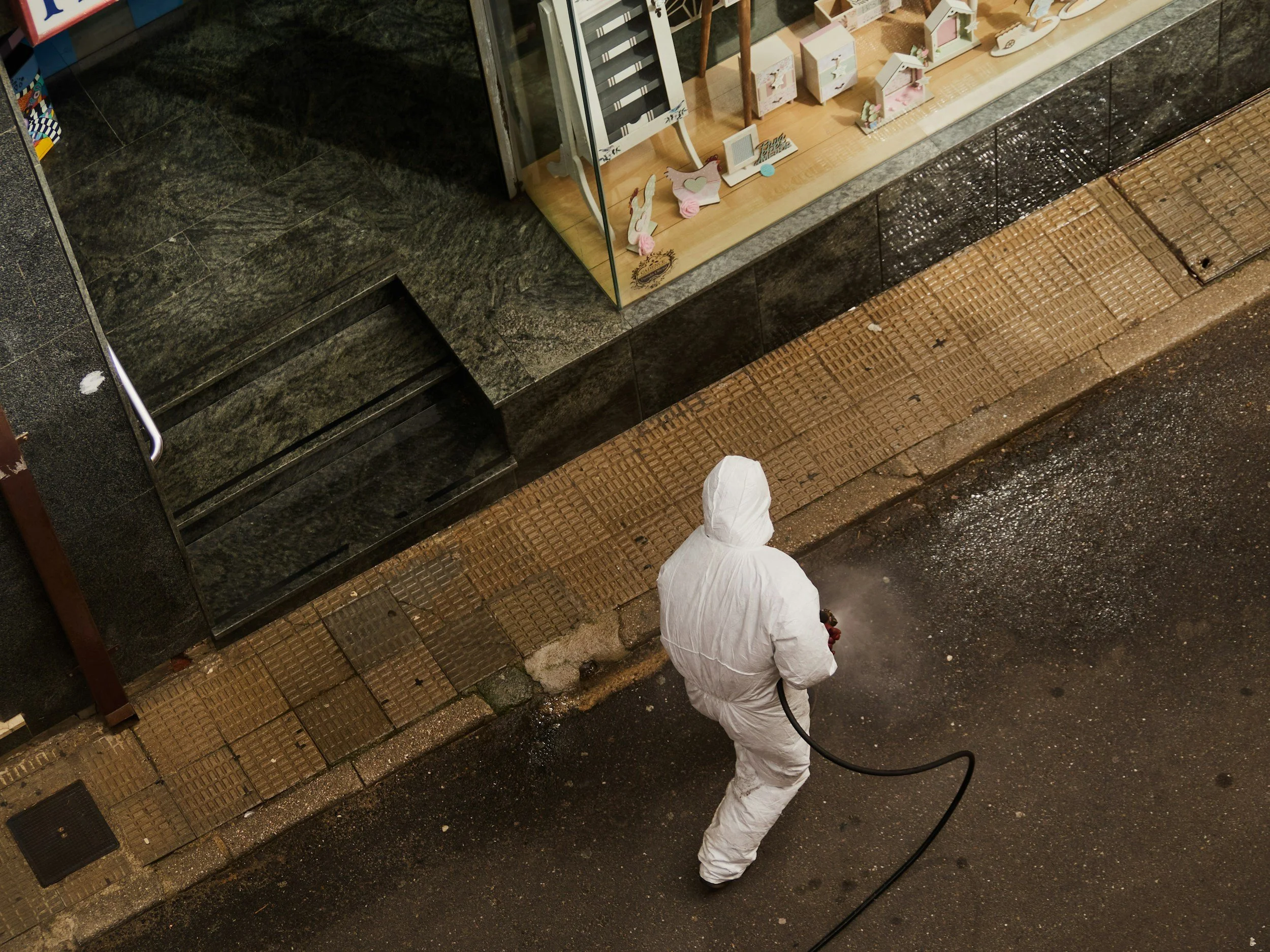 A person wearing a white protective suit and holding a hose sprays a storefront window display at night. The display contains decorative items, and the scene appears to be outside on a city street with wet pavement.