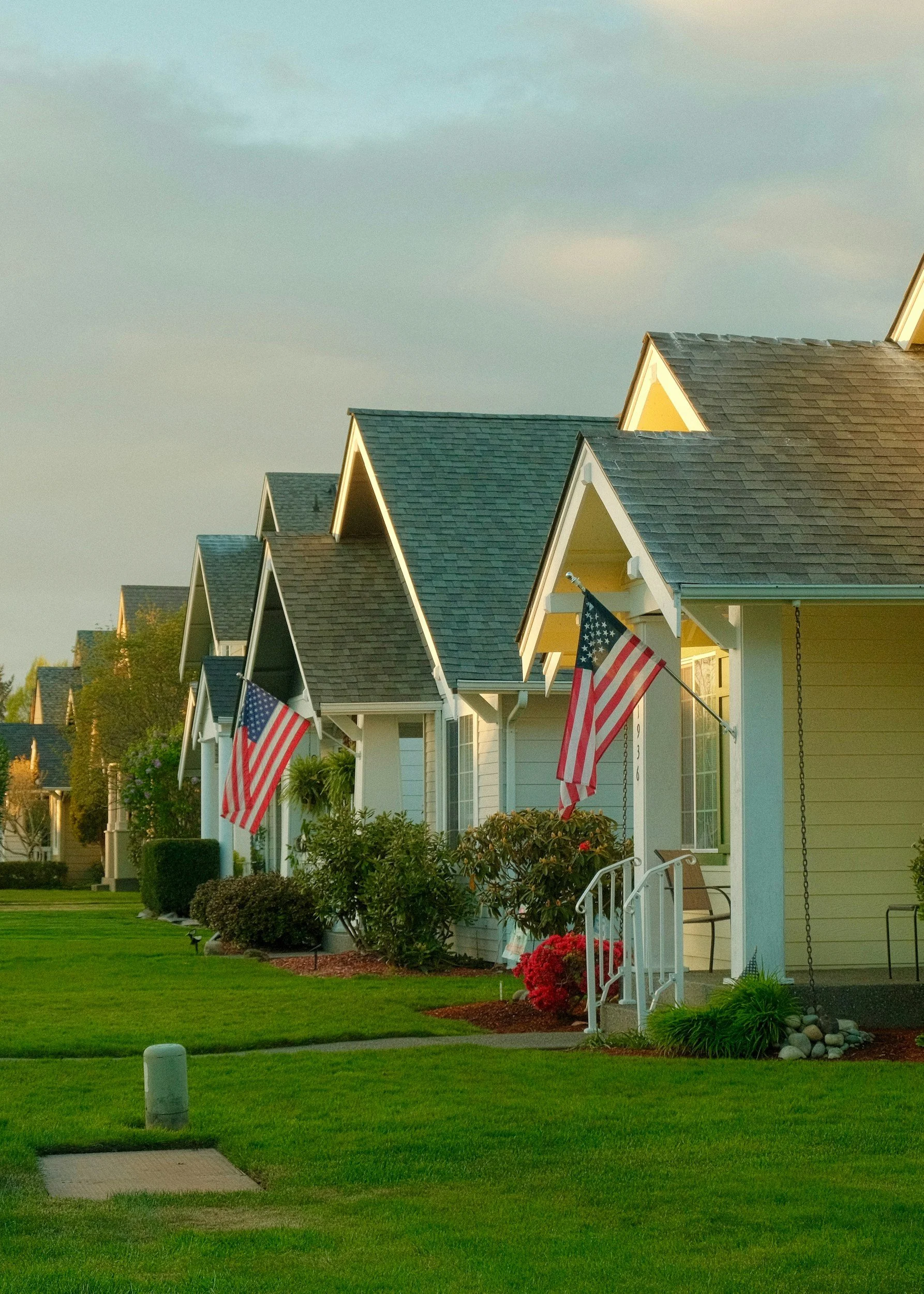 Row of yellow houses with front porches, American flags, well-maintained lawns, flower bushes, and a sidewalk in a suburban neighborhood, captured in late afternoon sunlight.