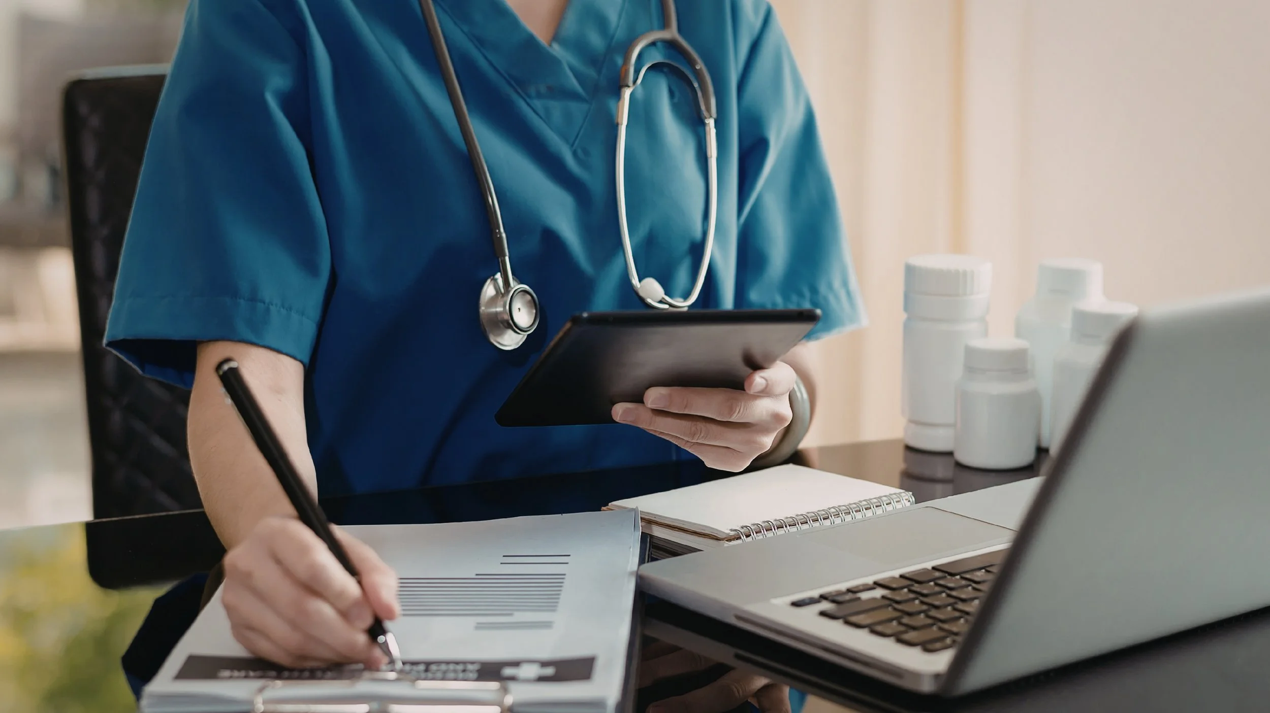 Medical professional in blue scrubs with stethoscope around neck using a tablet, taking notes on a clipboard, with medication bottles, a laptop, and a notebook on the desk.