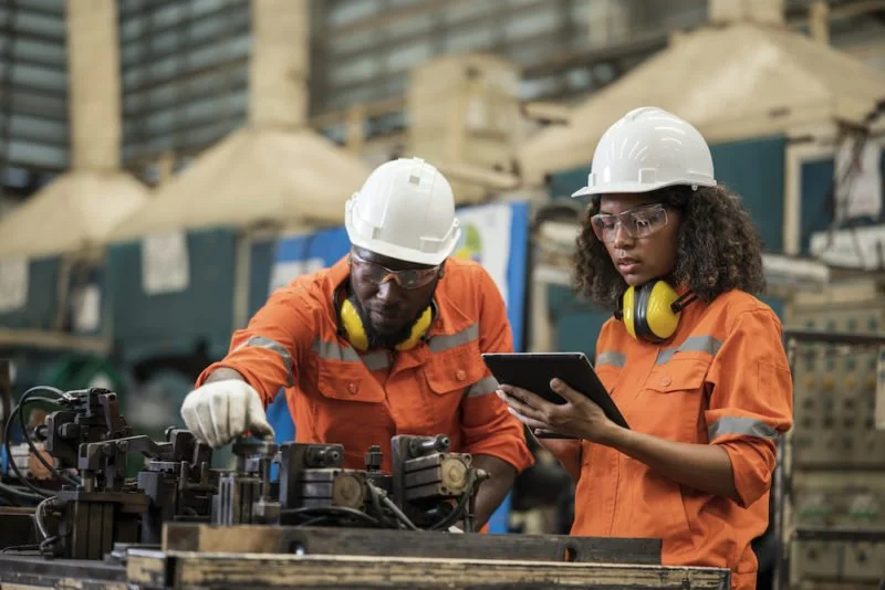 Two factory workers, a man and a woman, wearing orange uniforms, white safety helmets, ear protection, and safety glasses, are working on machinery at an industrial site.