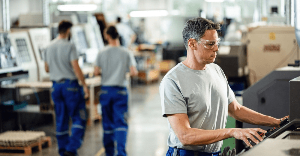 A woman working on a control panel in a manufacturing plant with other workers in the background.