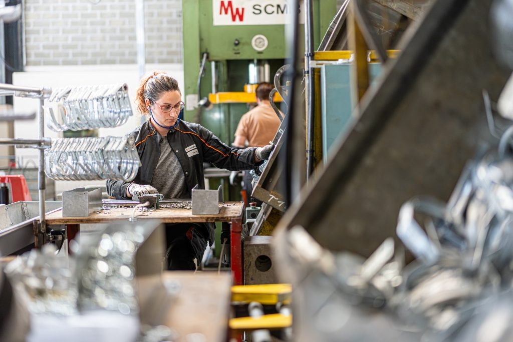 A woman wearing glasses, gloves, and a black jacket works in a factory with machinery and metal parts. She is focused on her task at a workbench.