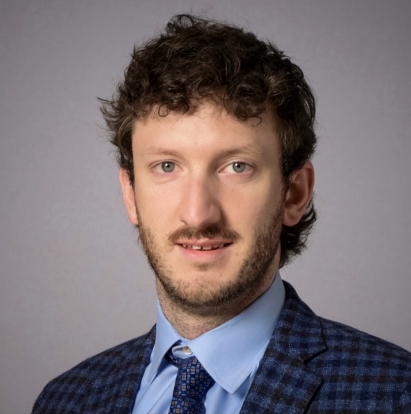 A man with wavy brown hair and a beard wearing a checkered blue suit jacket, light blue shirt, and patterned tie, looking at the camera against a plain gray background.