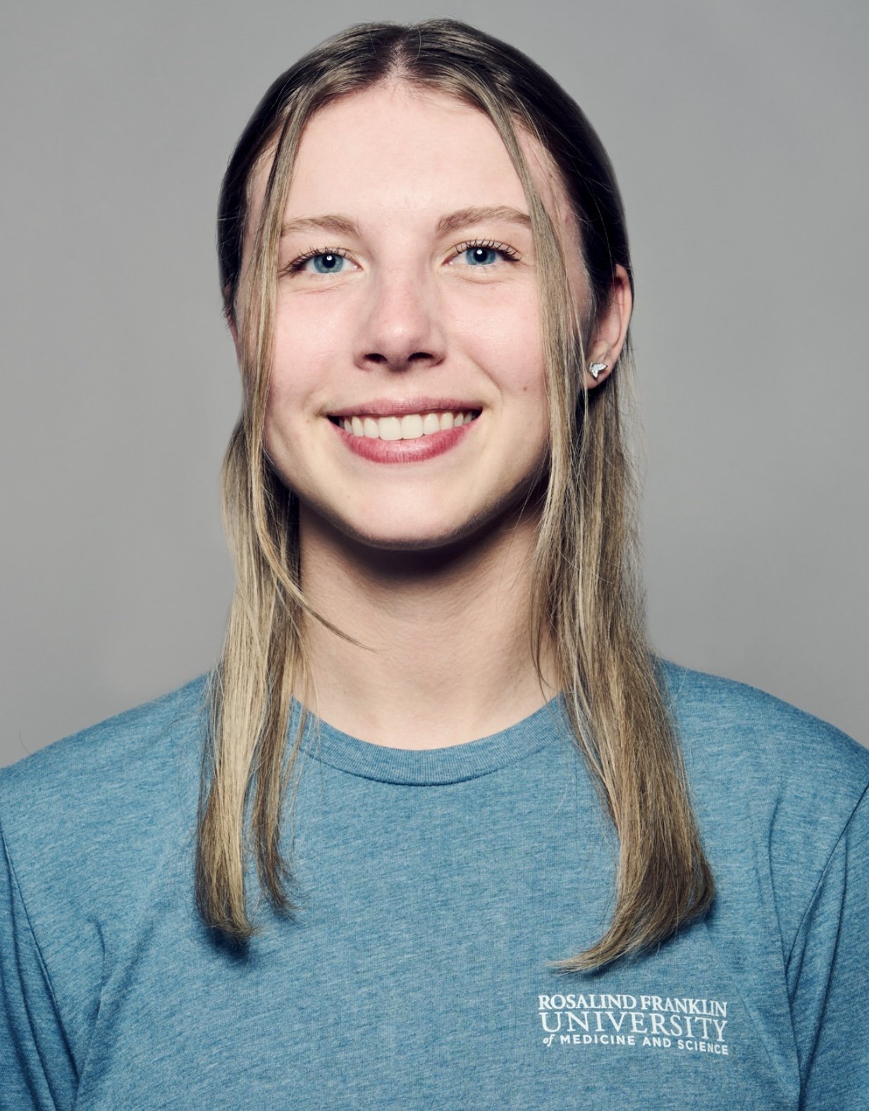 Rachel Ludwig. Smiling young woman with long blonde hair, blue eyes, wearing a blue Rosalind Franklin University T-shirt against a gray background.