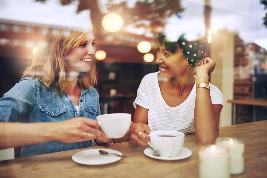 Two women smiling and chatting at a café, with coffee cups on the table, one woman holding a bowl.