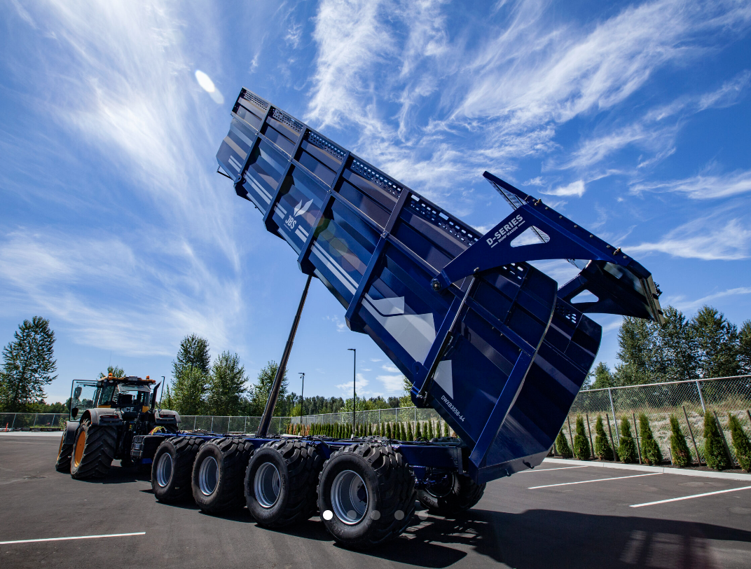 A large dump trailer attached to a tractor is raised at an angle, revealing its blue color and large wheels, set in a parking lot with trees and a blue sky with clouds in the background.