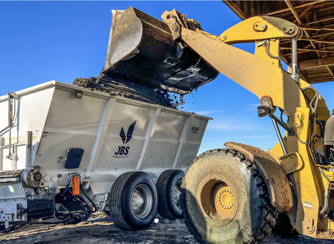 A yellow front loader excavator unloading dirt into a white dump trailer on a construction site, with a clear blue sky in the background.