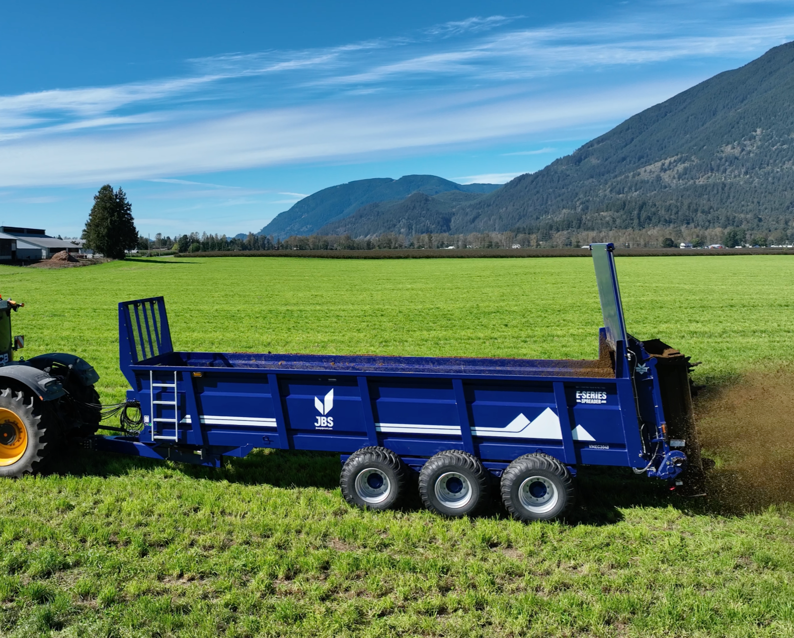 Blue agricultural trailer attached to a tractor in a green field with mountains and blue sky in the background.