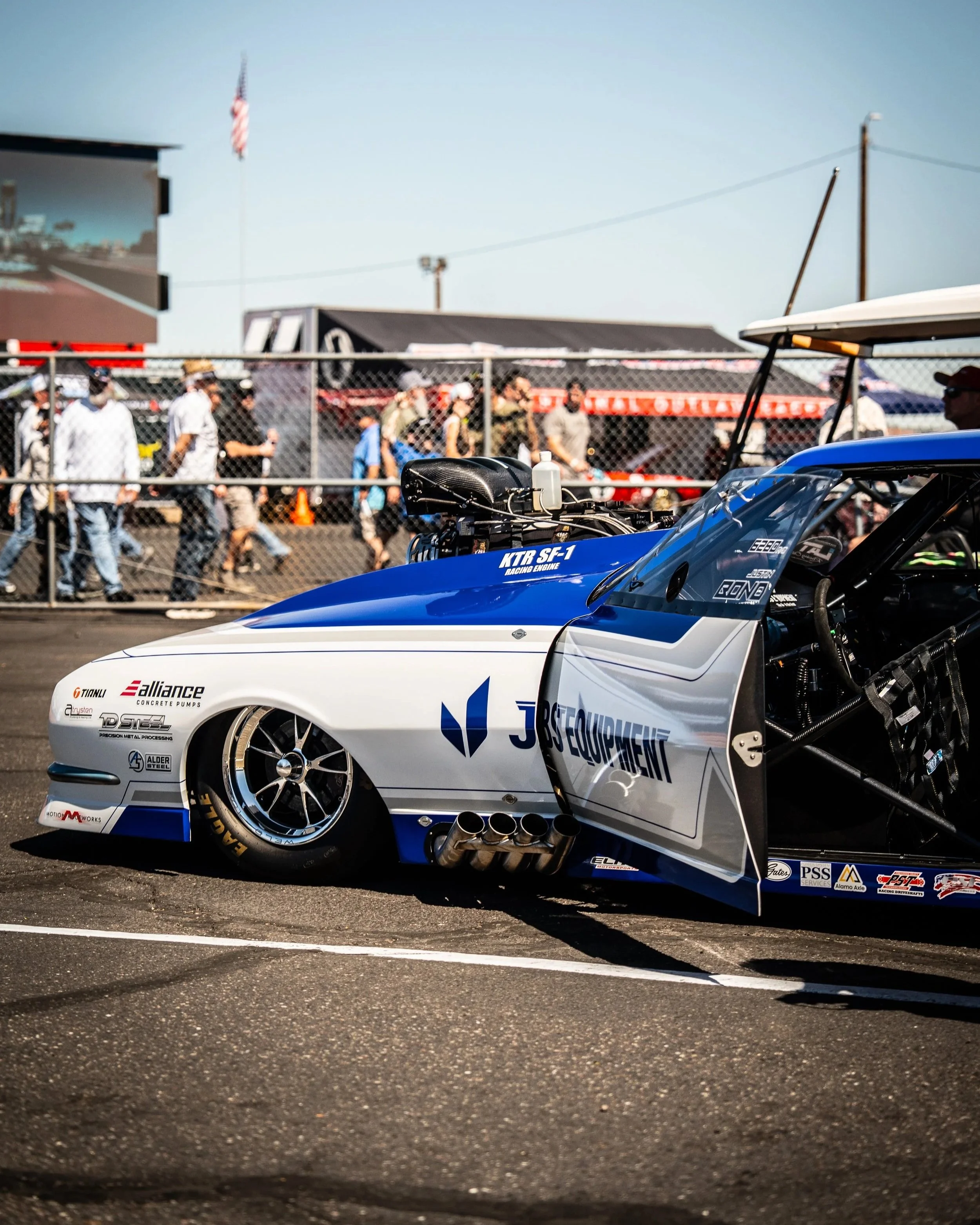 Close-up of a race car with sponsorship logos, parked on a race track, with people in the background behind a chain-link fence and racing tents under a clear sky.